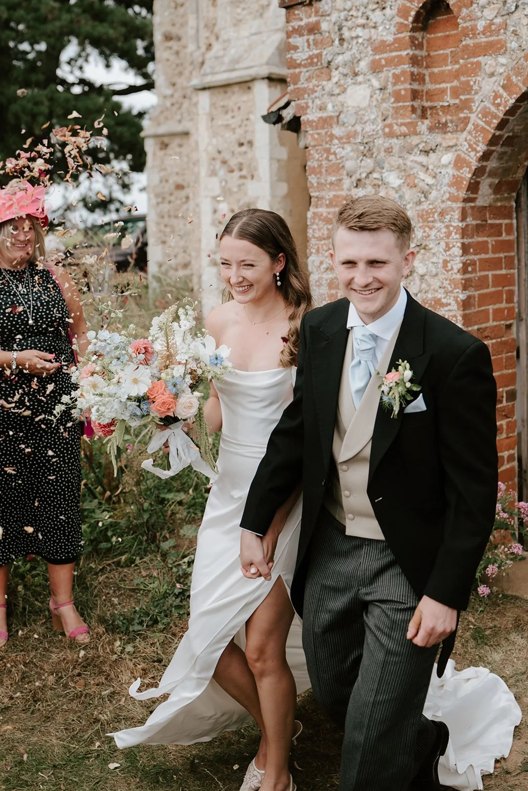 Happy bride and groom holding hands and smiling, walking outdoors near a brick and stone building, with wedding guests around, during a wedding celebration.