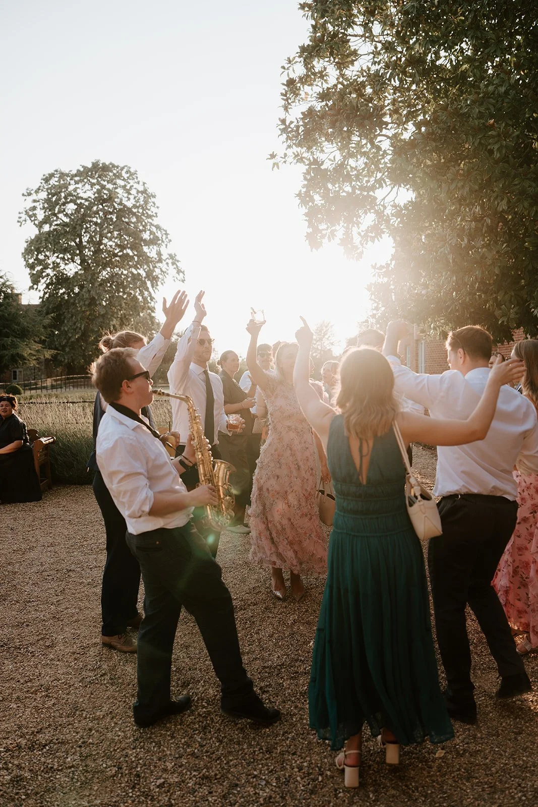 People dancing and celebrating outdoors in the evening sunlight, some holding drinks, with trees and a building in the background.