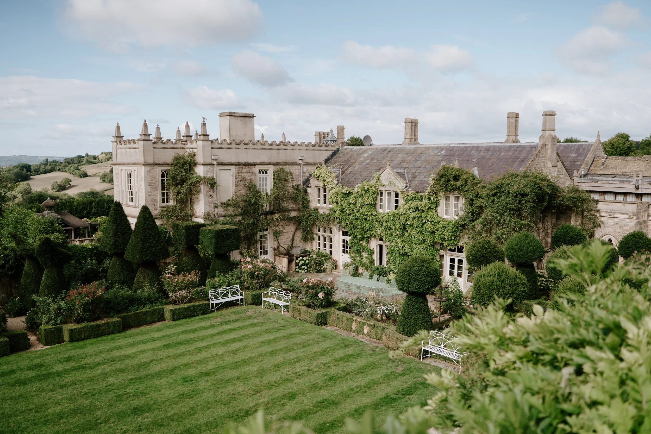 A large historic stone mansion with ivy growing on the walls, surrounded by a well-maintained garden with benches, trimmed hedges, and flower beds under a partly cloudy sky.