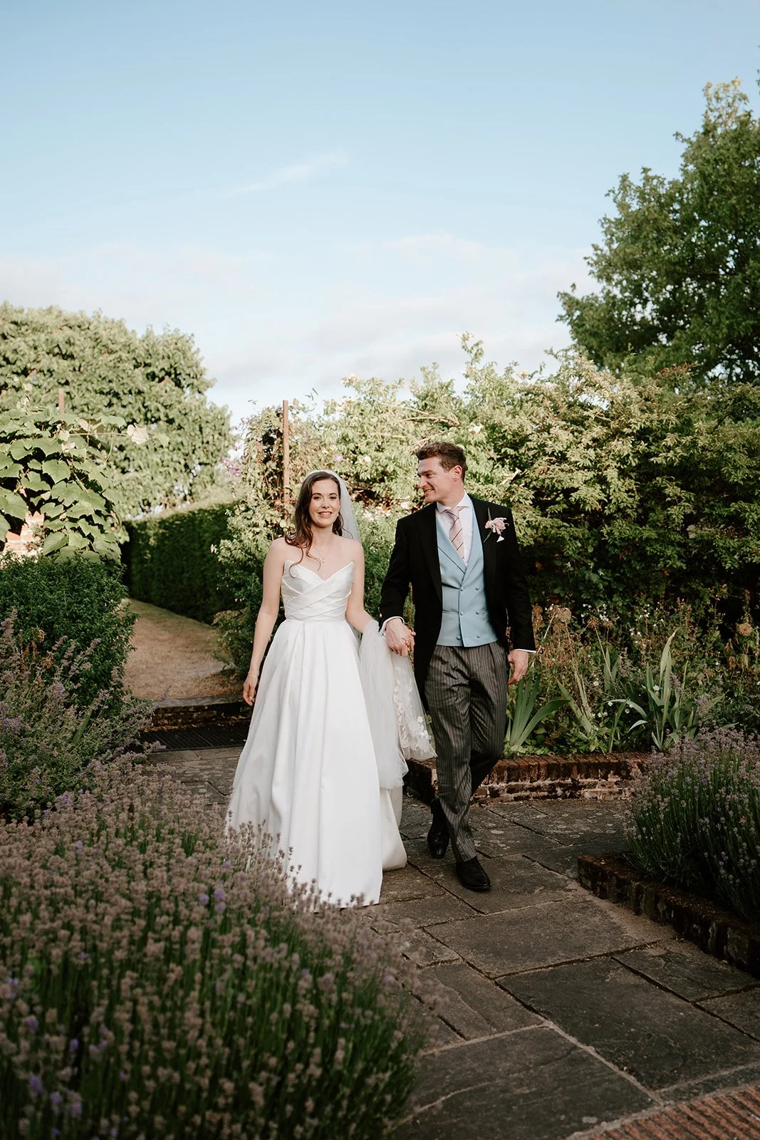 Bride and groom walking hand in hand outdoors in a garden on their wedding day, surrounded by green shrubs and flowering plants under a clear blue sky.