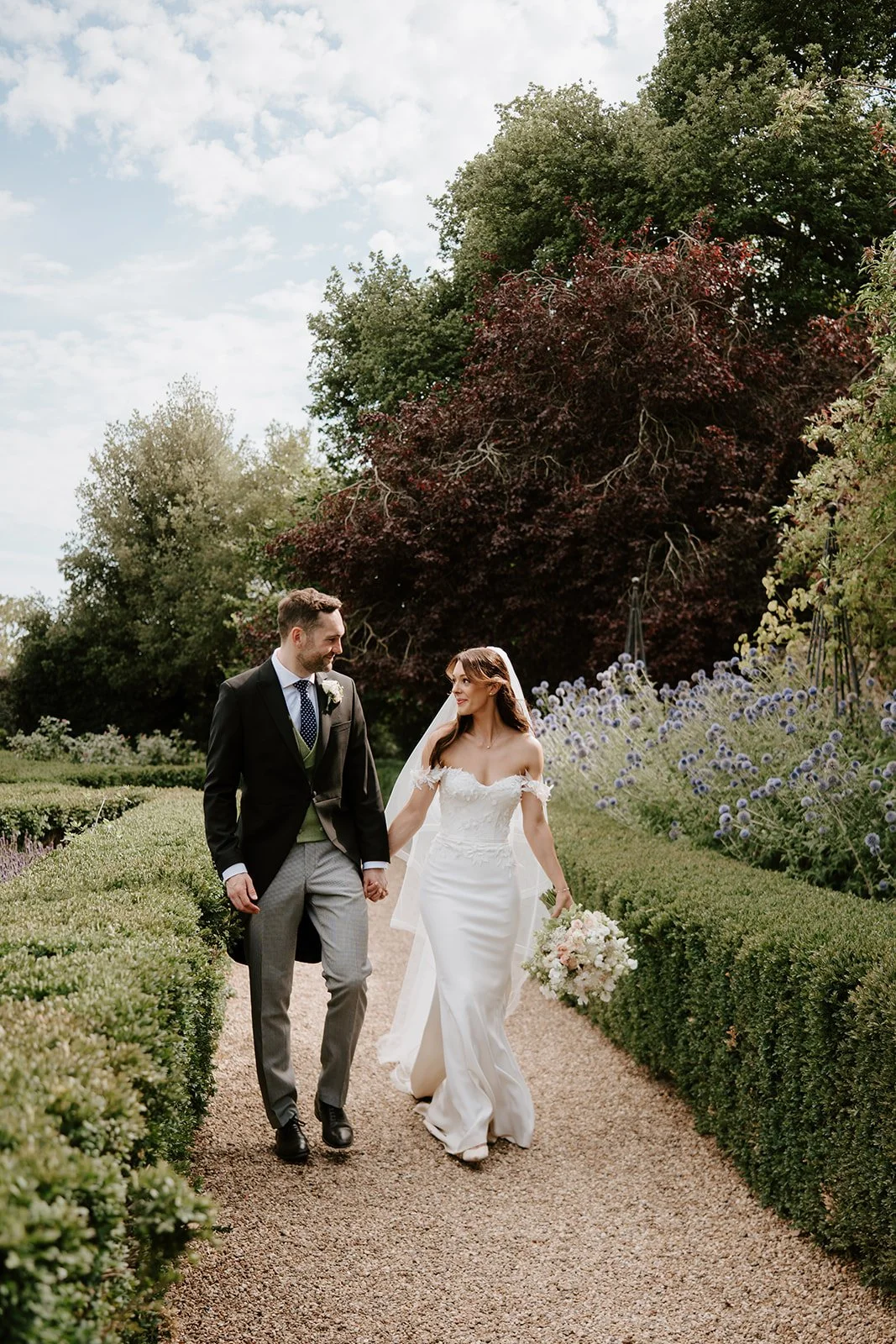 A bride and groom walking hand in hand along a garden path surrounded by lush greenery and blooming flowers on a sunny day.
