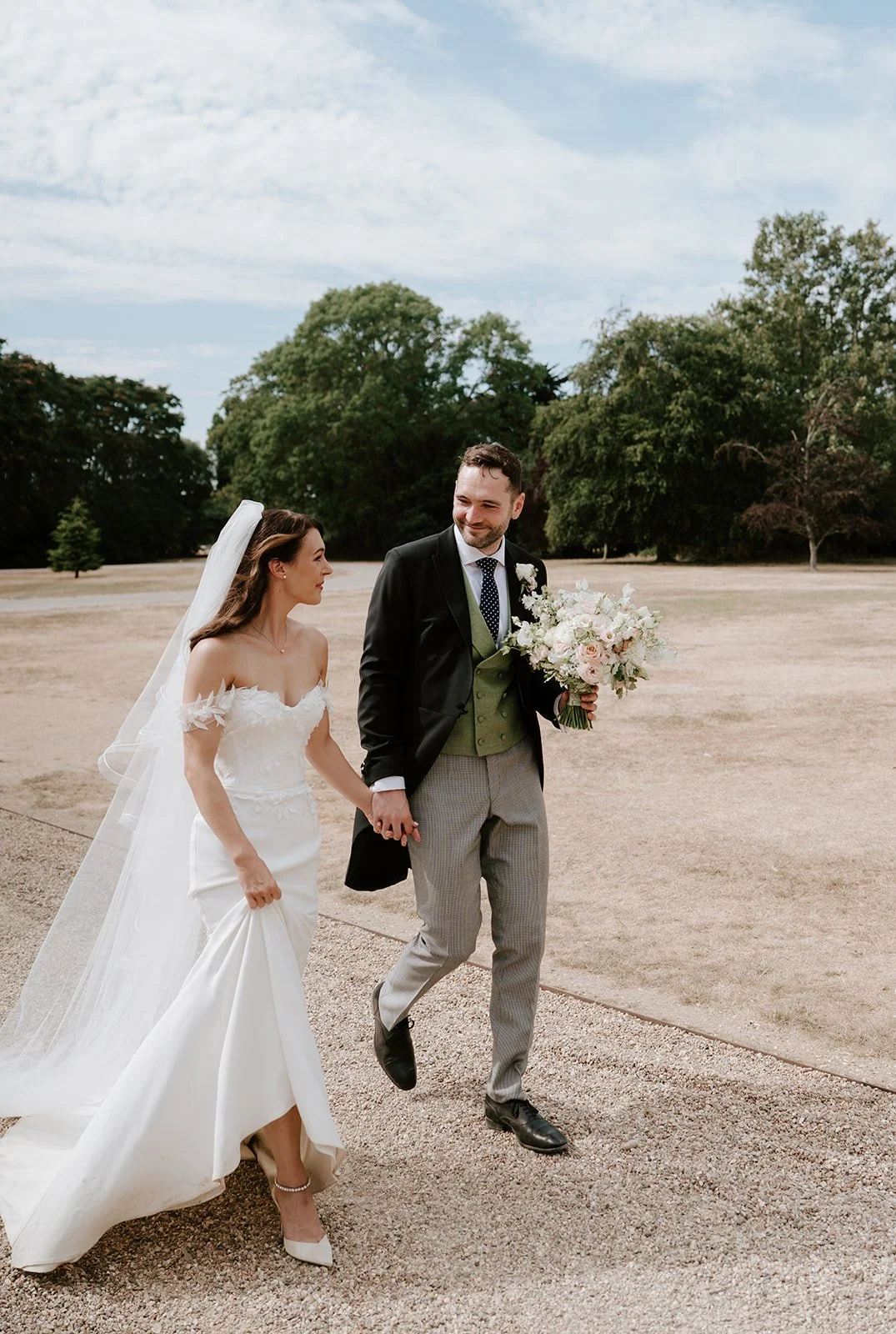 Bride and groom holding hands while walking outdoors on their wedding day. The bride is wearing a strapless white gown and veil, and the groom is dressed in a black jacket with a green vest and gray plaid pants, holding a bouquet of flowers. Trees an