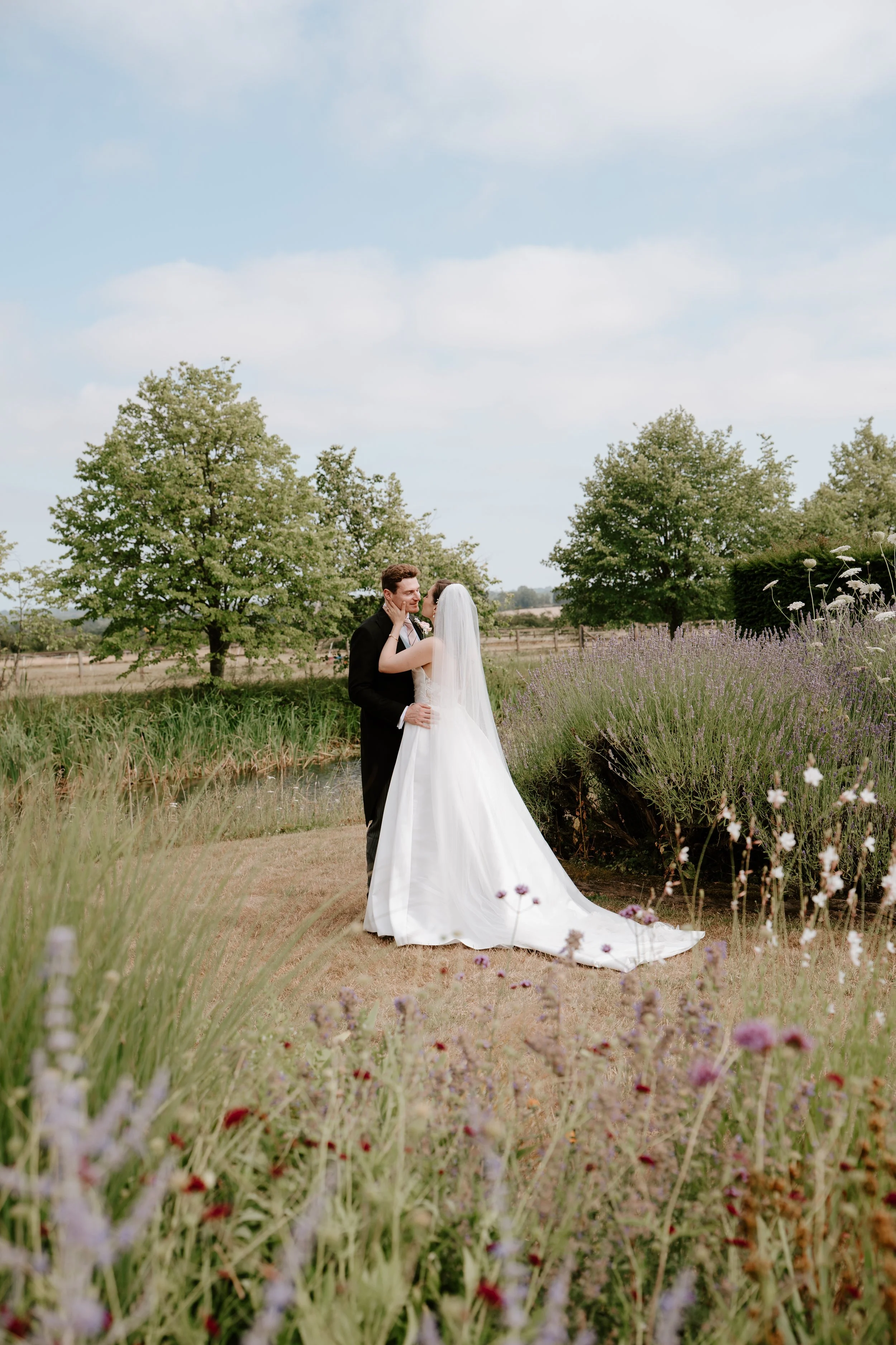A bride and groom sharing a romantic moment outdoors, with trees and purple flowers surrounding them on a sunny day.