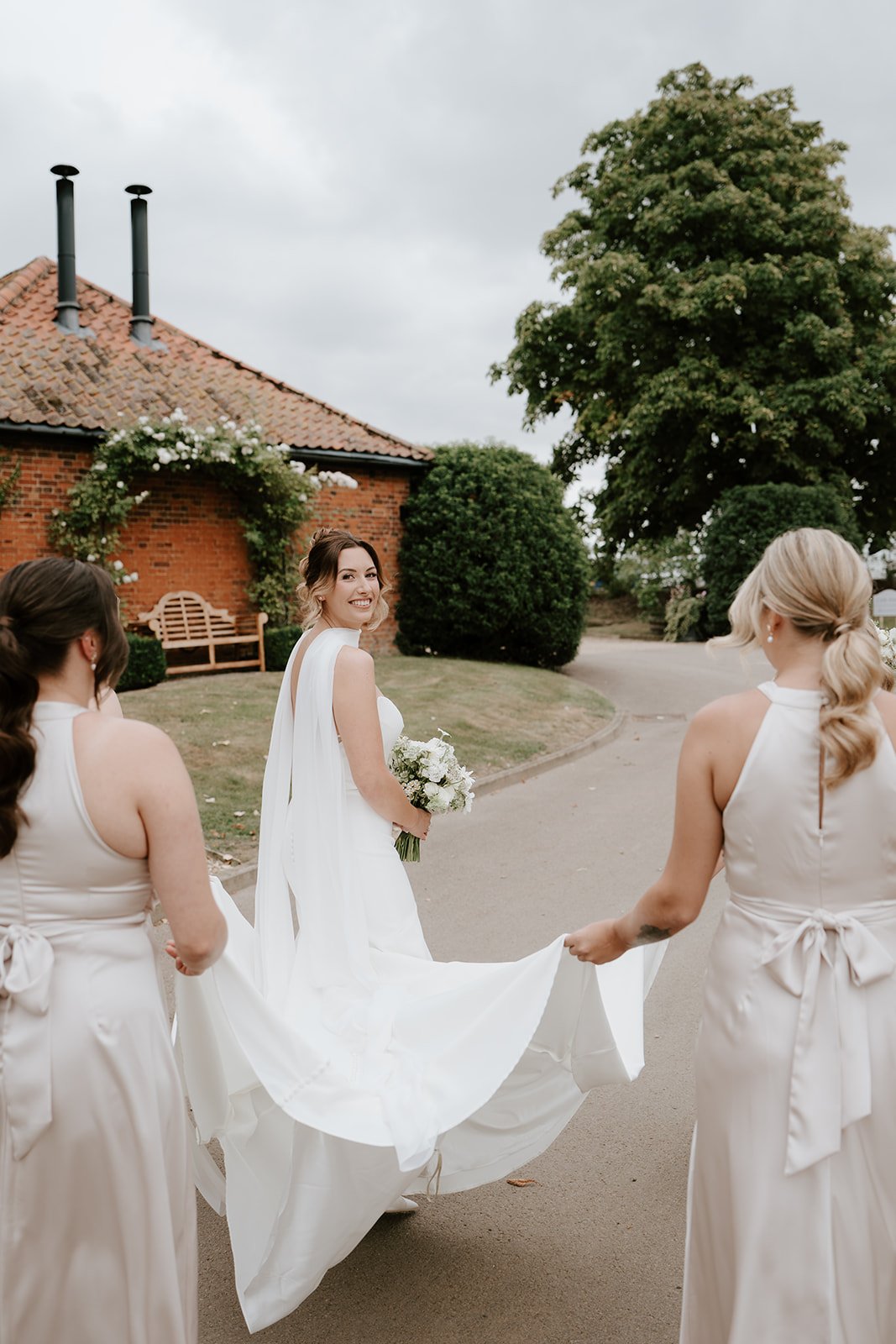 A bride smiling and holding a bouquet of white flowers, walking on a suburban street with two bridesmaids holding the train of her dress on each side. The background features a brick house with a tiled roof and chimney, a wooden bench, and a large leafy tree.