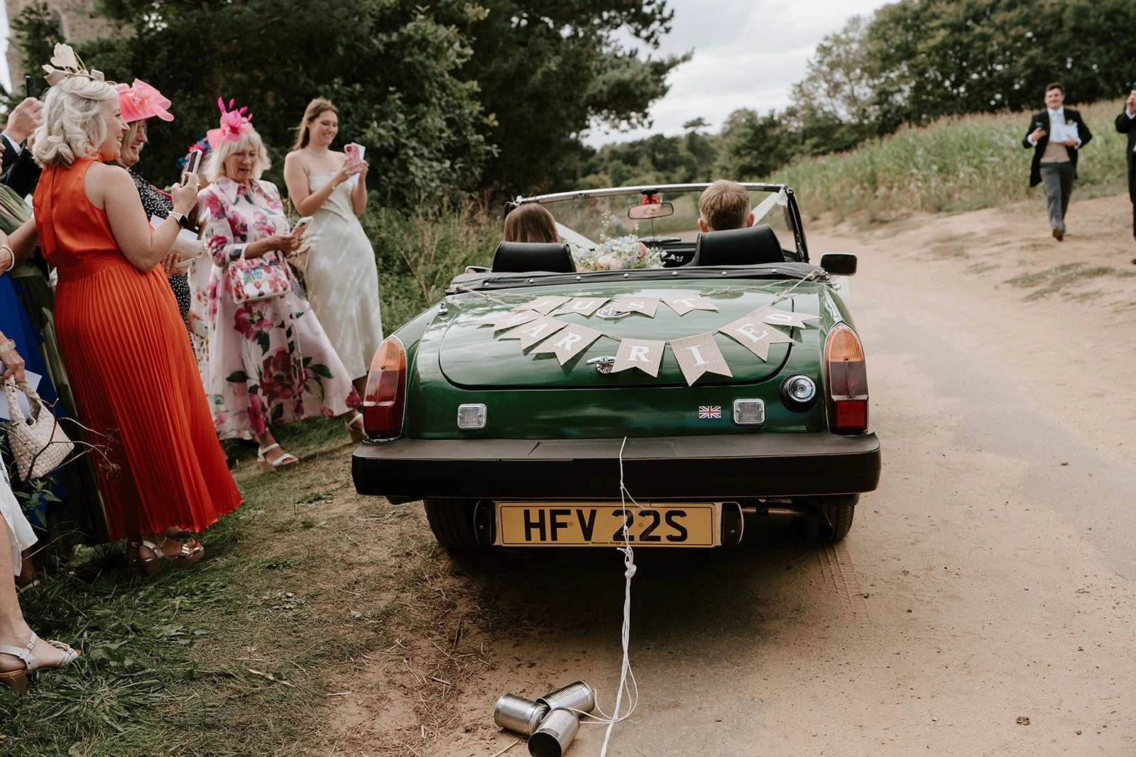 A green convertible car decorated for a wedding with a "Just Married" banner, parked on a dirt road. People dressed in colorful attire, some wearing hats, are gathered nearby taking photos. The scene is outdoors with trees and open land in the background.
