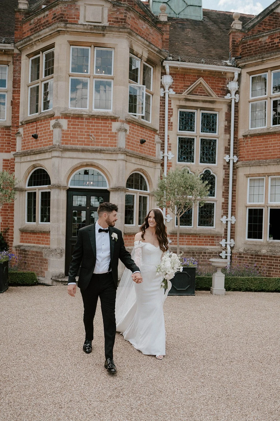 Bride and groom holding hands, bride wearing Suzanne Neville lace off shoulder dress