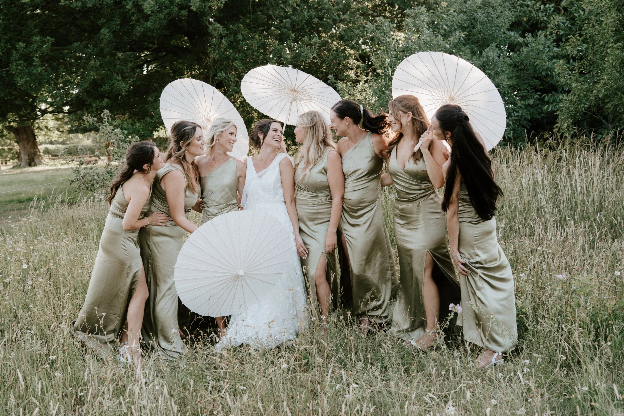 A group of women in matching gold satin dresses standing in a grassy field, holding white parasols, smiling and looking at each other, with large trees in the background.