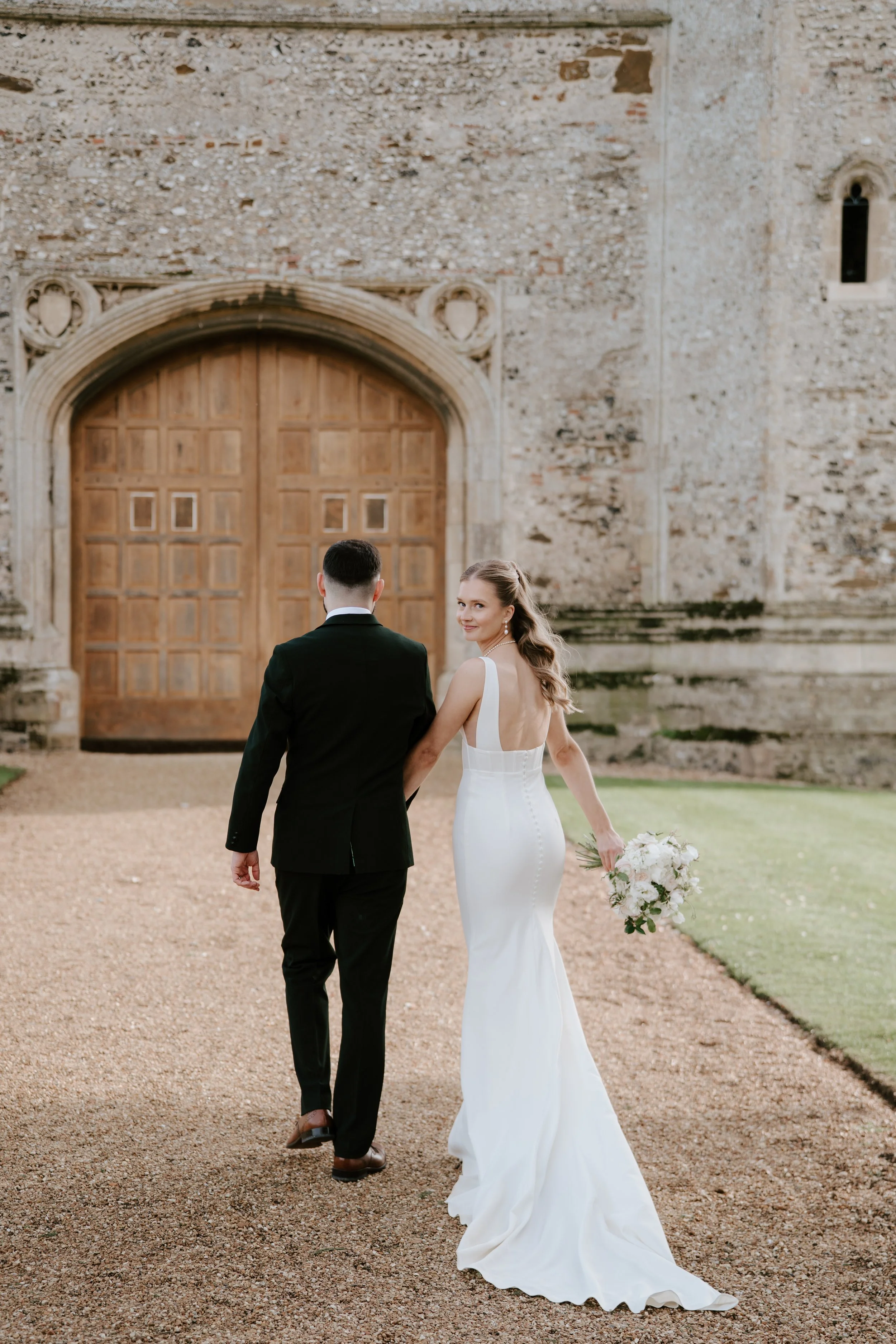 A bride and groom walking hand in hand outside a historic stone building, with the bride smiling and holding a bouquet of white flowers.