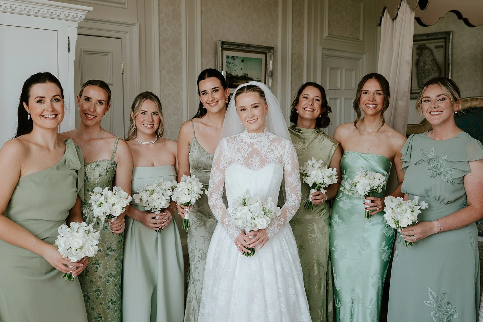 A bride and her eight bridesmaids standing together indoors, holding white bouquets, smiling for a photo.