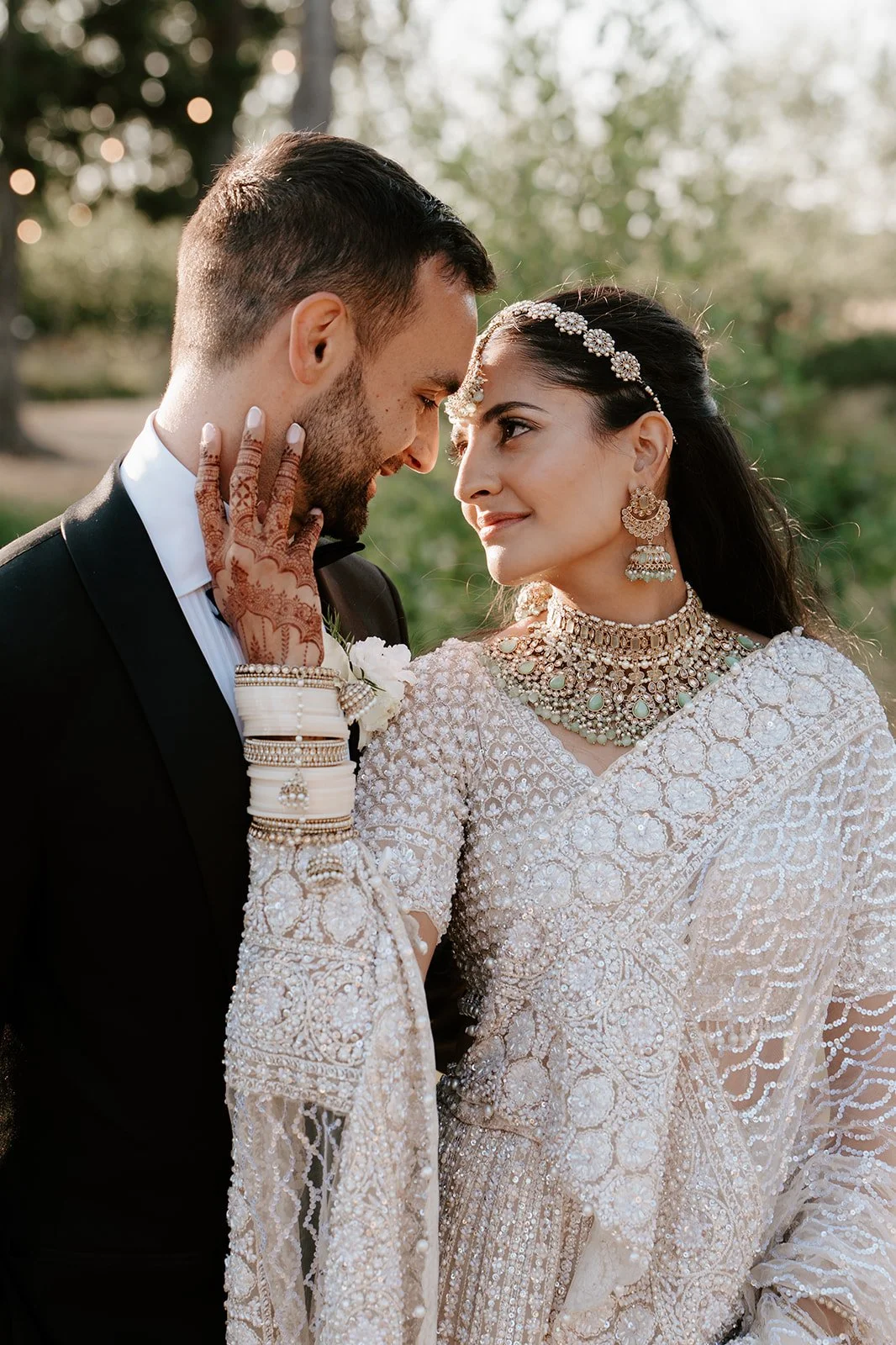 A bride and groom share a close moment outdoors, dressed in wedding attire with the bride wearing traditional jewelry and henna on her hand.