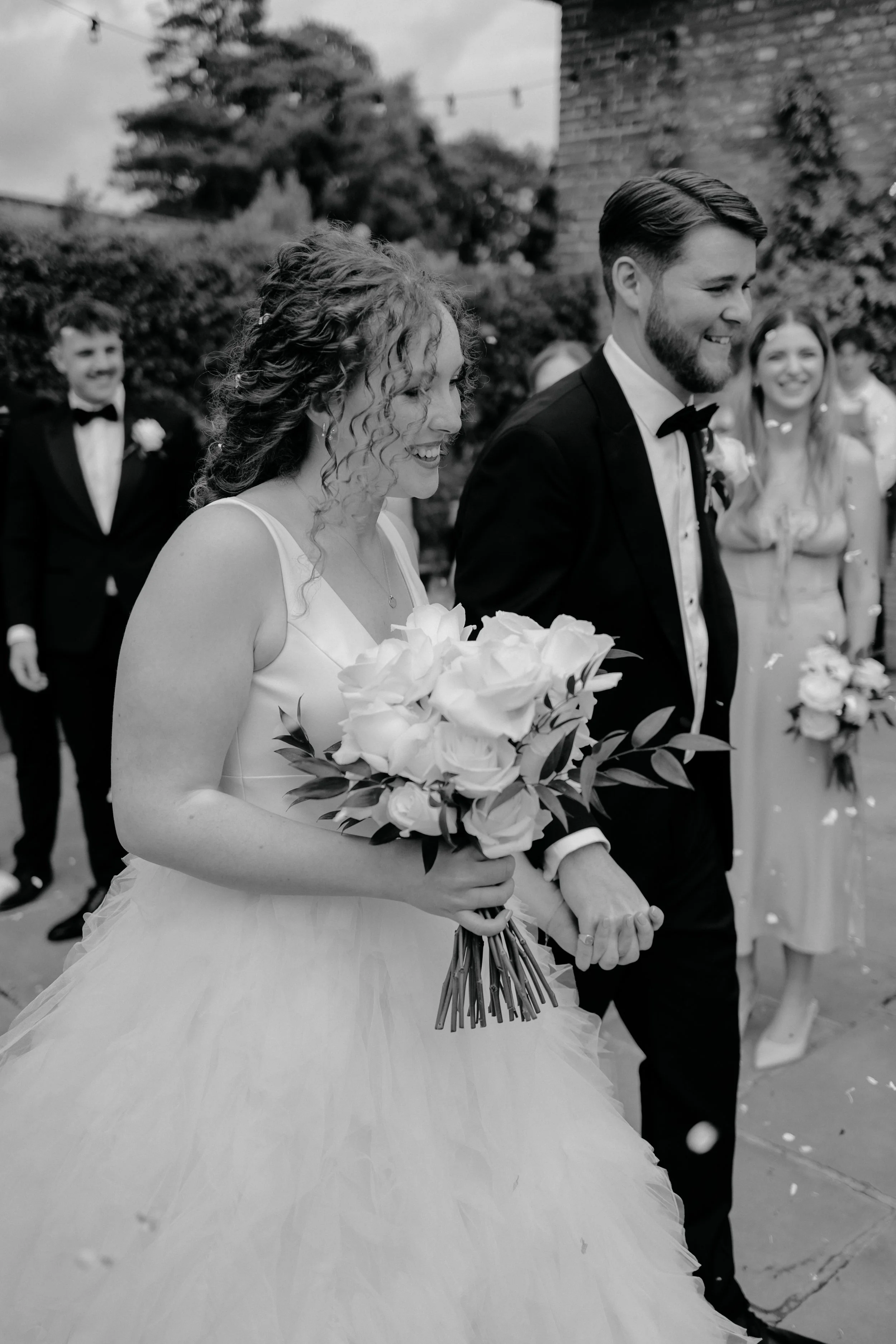 A bride and groom walking hand in hand during their wedding, smiling. The bride has curly hair and is holding a bouquet of roses. The groom has a beard and is wearing a tuxedo. Several guests in formal attire are visible in the background.