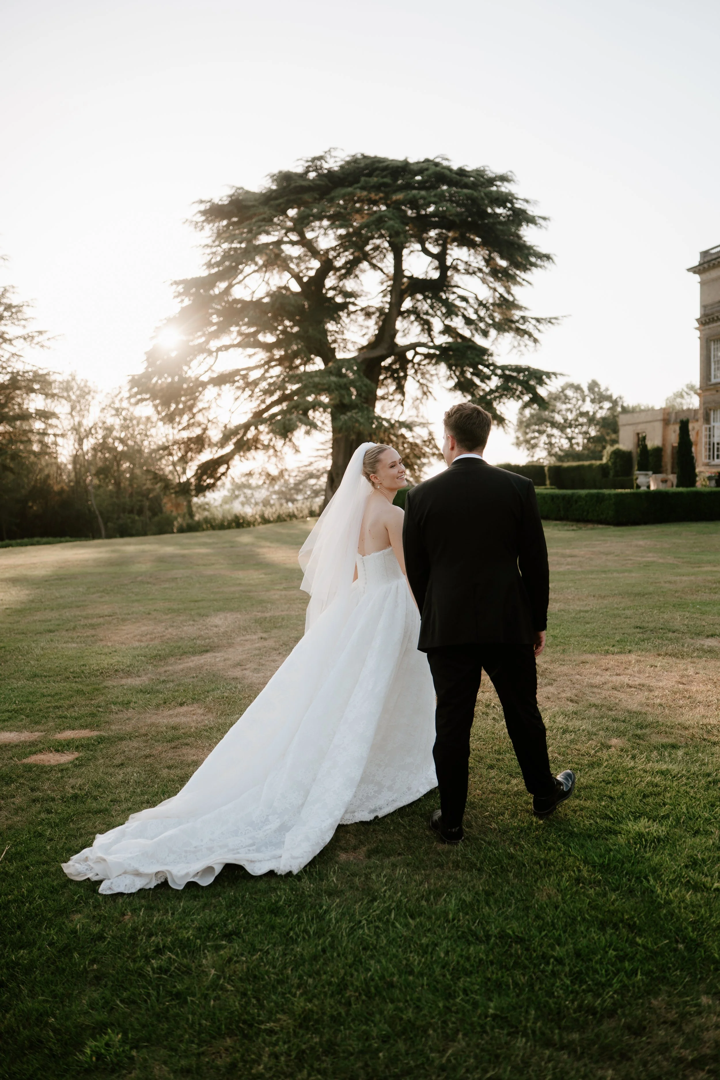 A bride and groom walking on grass lawn in a garden setting during sunset, with the bride in a white wedding gown and veil, and the groom in a black suit, facing each other and smiling.