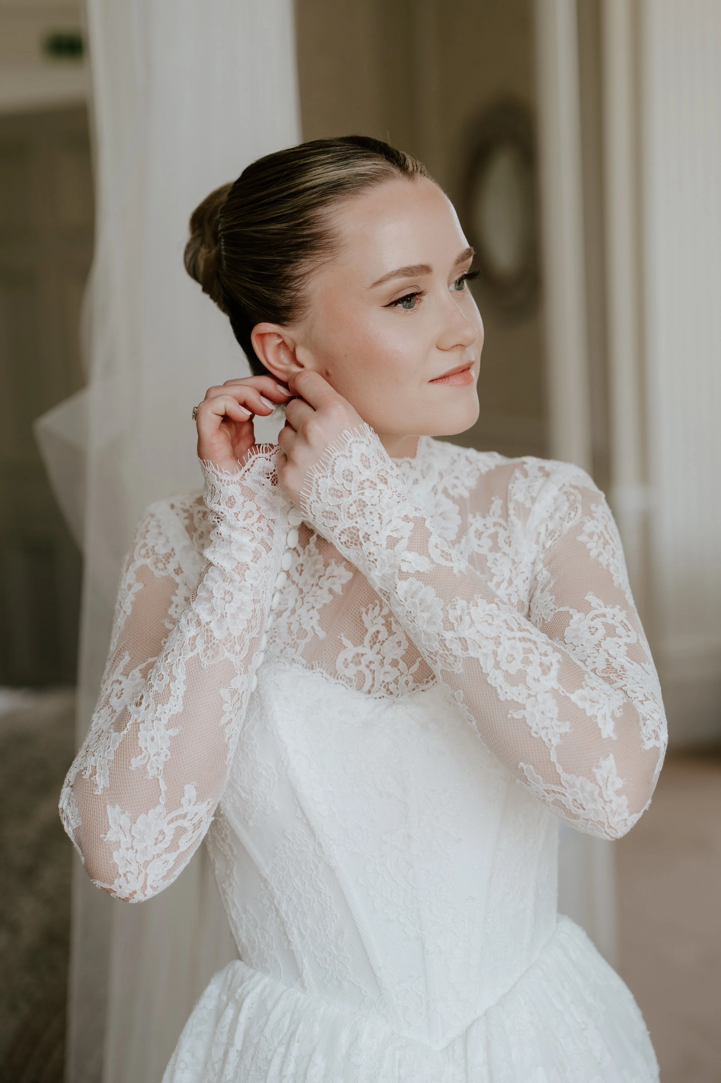 A woman in a white lace wedding dress putting on an earring, with her hair styled in an elegant updo.