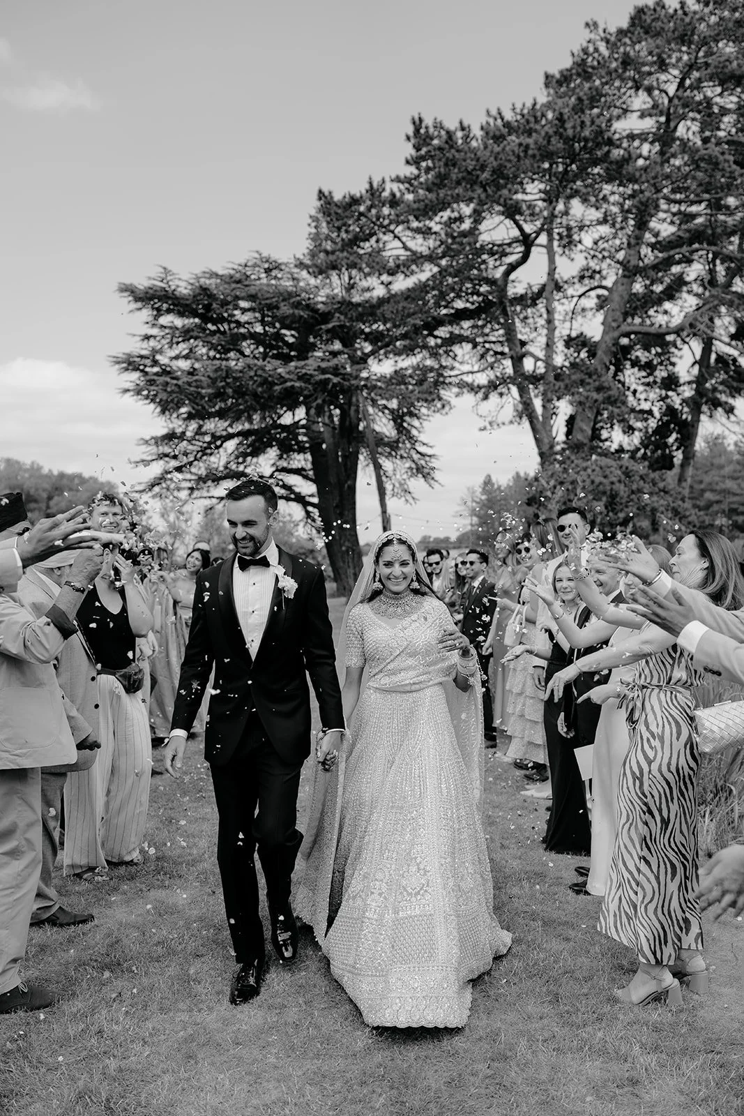 A black and white photo of a wedding celebration with a bride and groom walking hand in hand through a crowd of guests throwing confetti outdoors near trees.