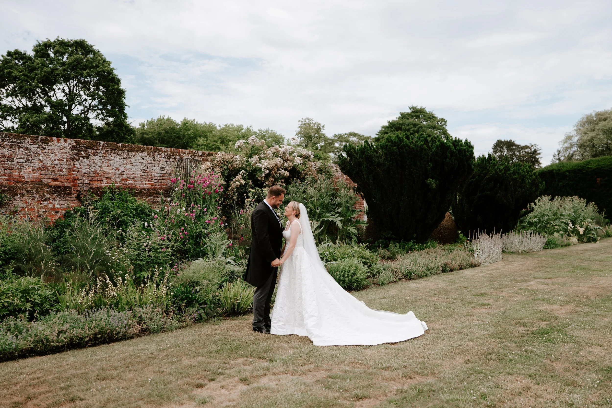 A bride and groom stand closely together in a garden, holding hands and smiling at each other, with a brick wall, blooming flowers, and trees in the background.