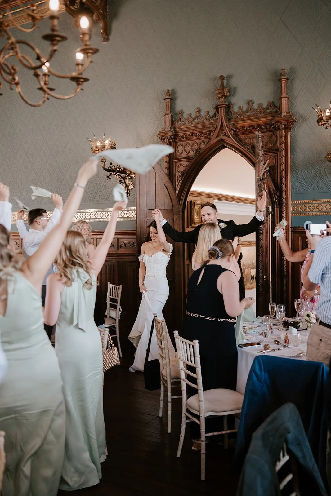 Bride and groom celebrating at their wedding reception with guests cheering and raising napkins in a decorated elegant room.