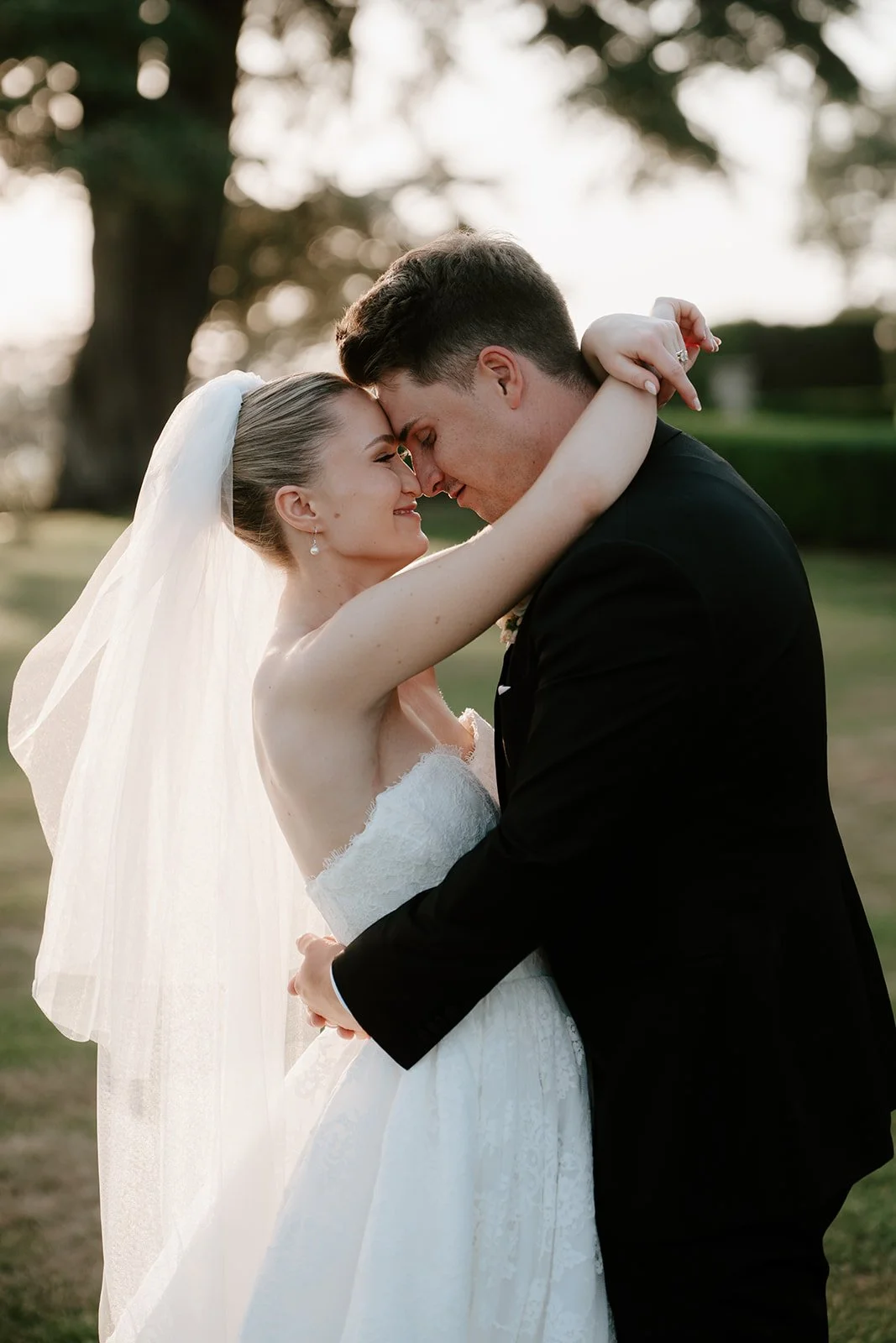 A bride and groom embracing outdoors during sunset, smiling with foreheads touching.