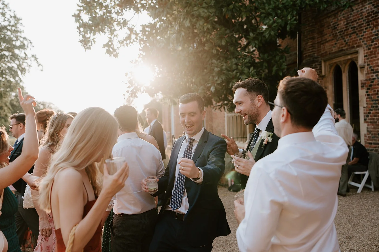 Group of friends celebrating at an outdoor wedding reception, smiling, dancing, and drinking together in warm sunlight.