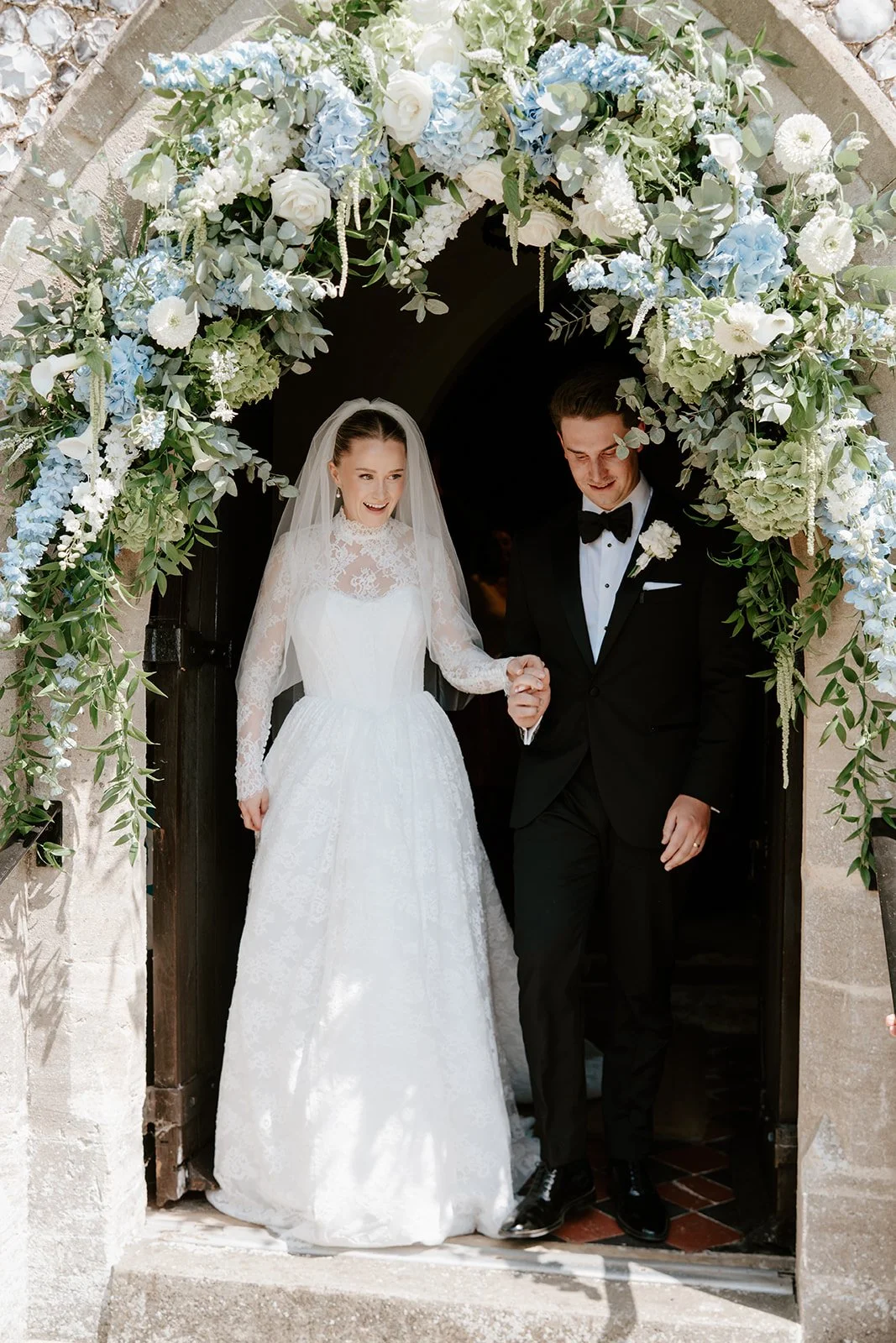 A bride and groom leaving a church under a floral arch, holding hands. The bride is wearing a white lace wedding dress with long sleeves and a veil, and the groom is in a black tuxedo with a bow tie. The floral arrangement above them includes white, 