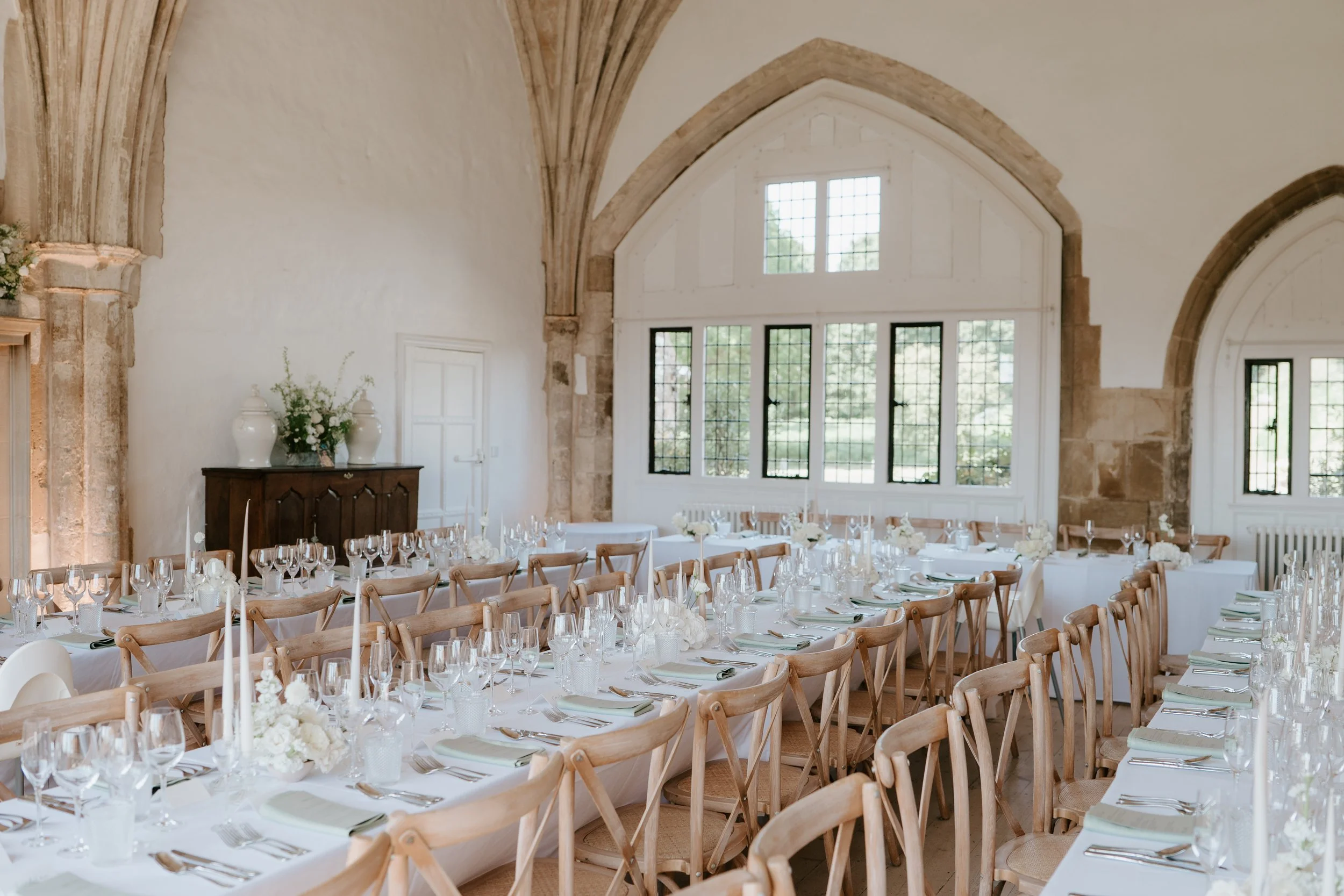 Elegant indoor wedding reception with round tables set with white tablecloths, wine glasses, silverware, and white floral centerpieces, featuring large arched windows and stone architectural details.