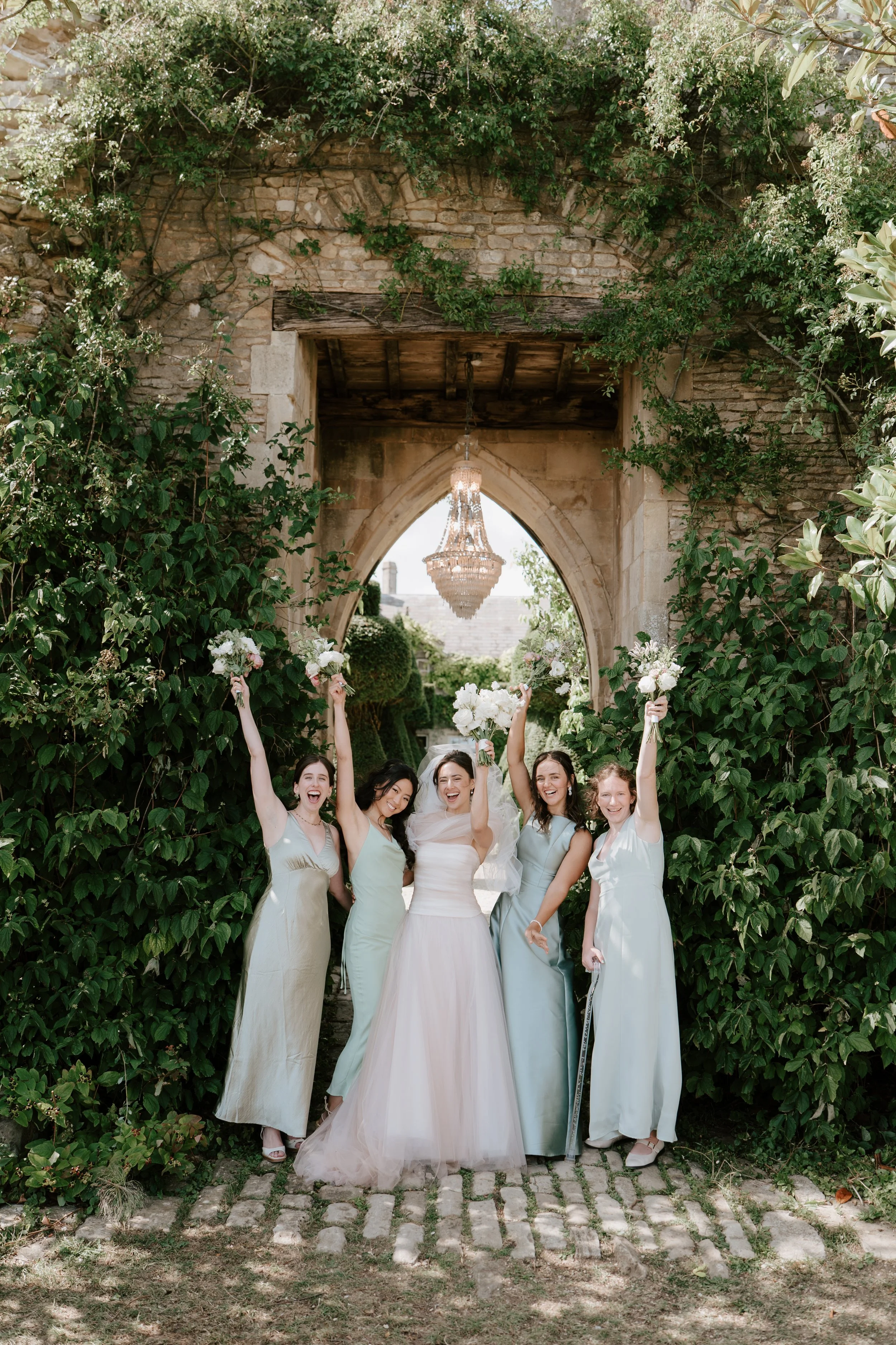 Six women in pastel-colored dresses celebrating outdoors under a stone archway with greenery, holding bouquets and smiling.
