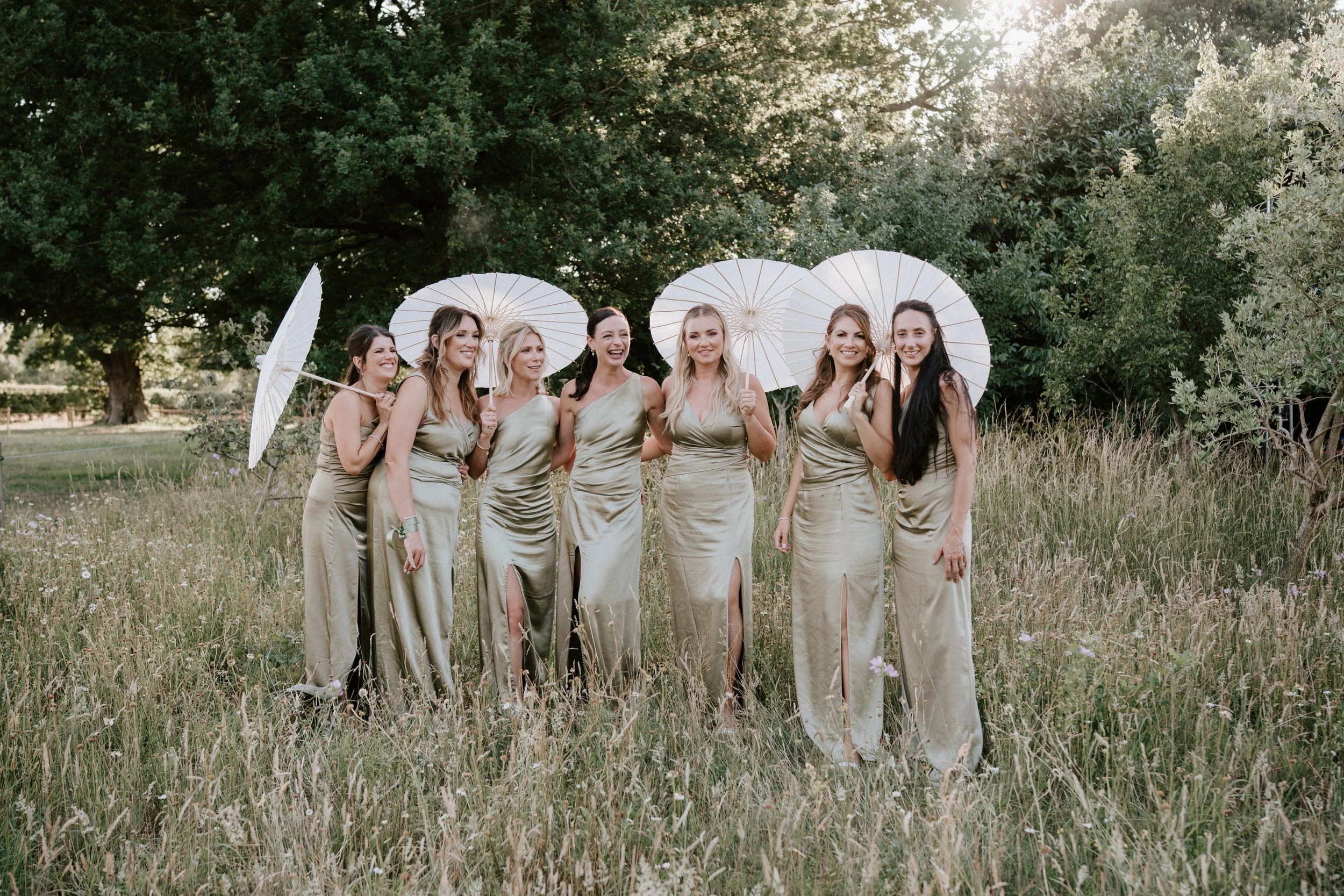 Group of seven women in gold satin dresses holding parasols in a grassy field with trees in the background.