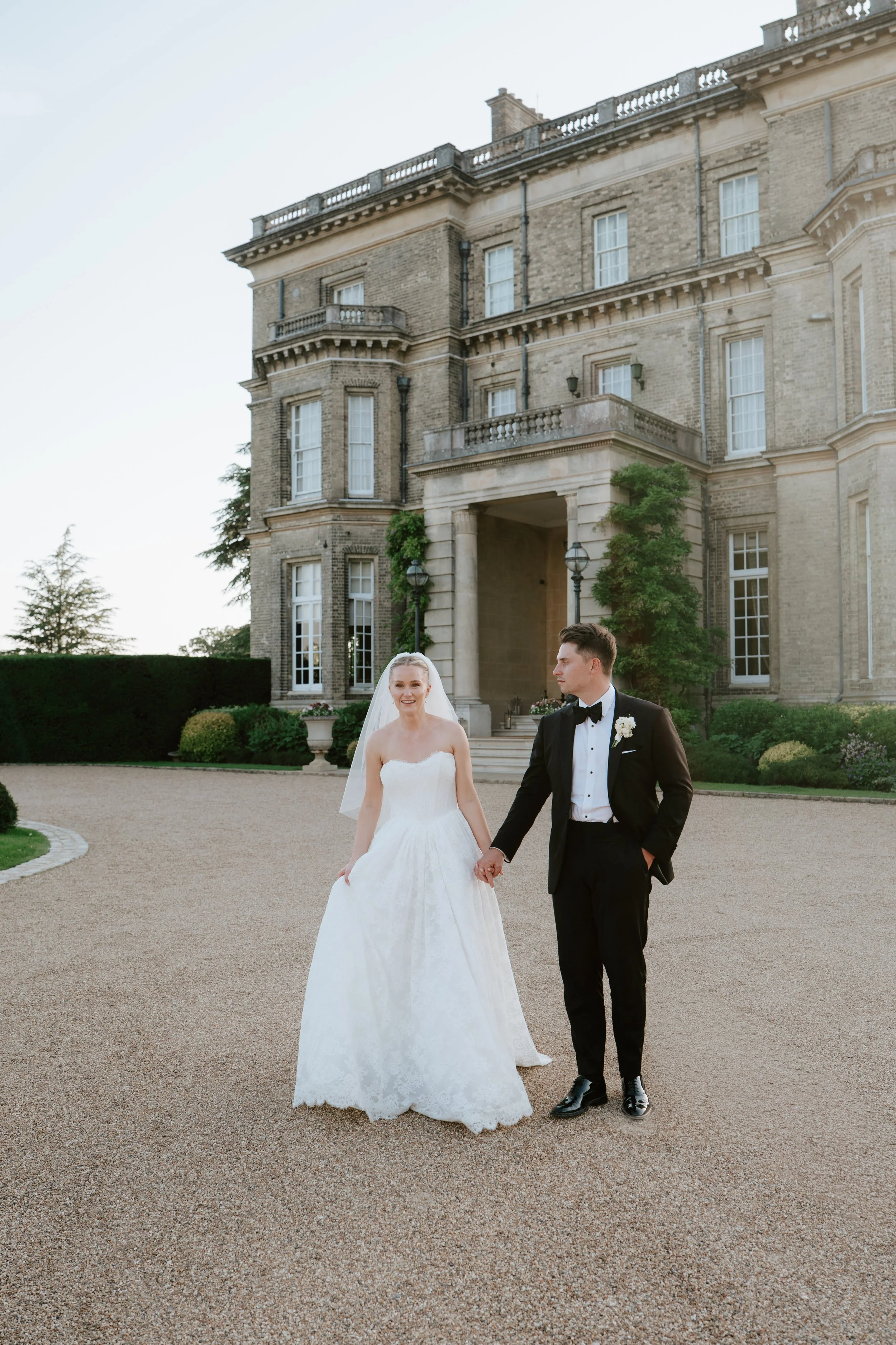 A bride and groom walking hand-in-hand outside a large, historic brick mansion during their wedding day.