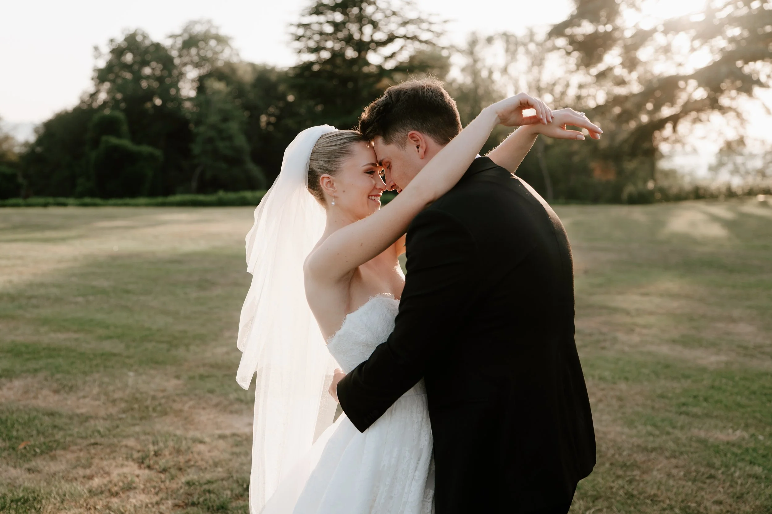 A bride and groom embracing outdoors during sunset, smiling with foreheads touching, with trees and open field in the background.
