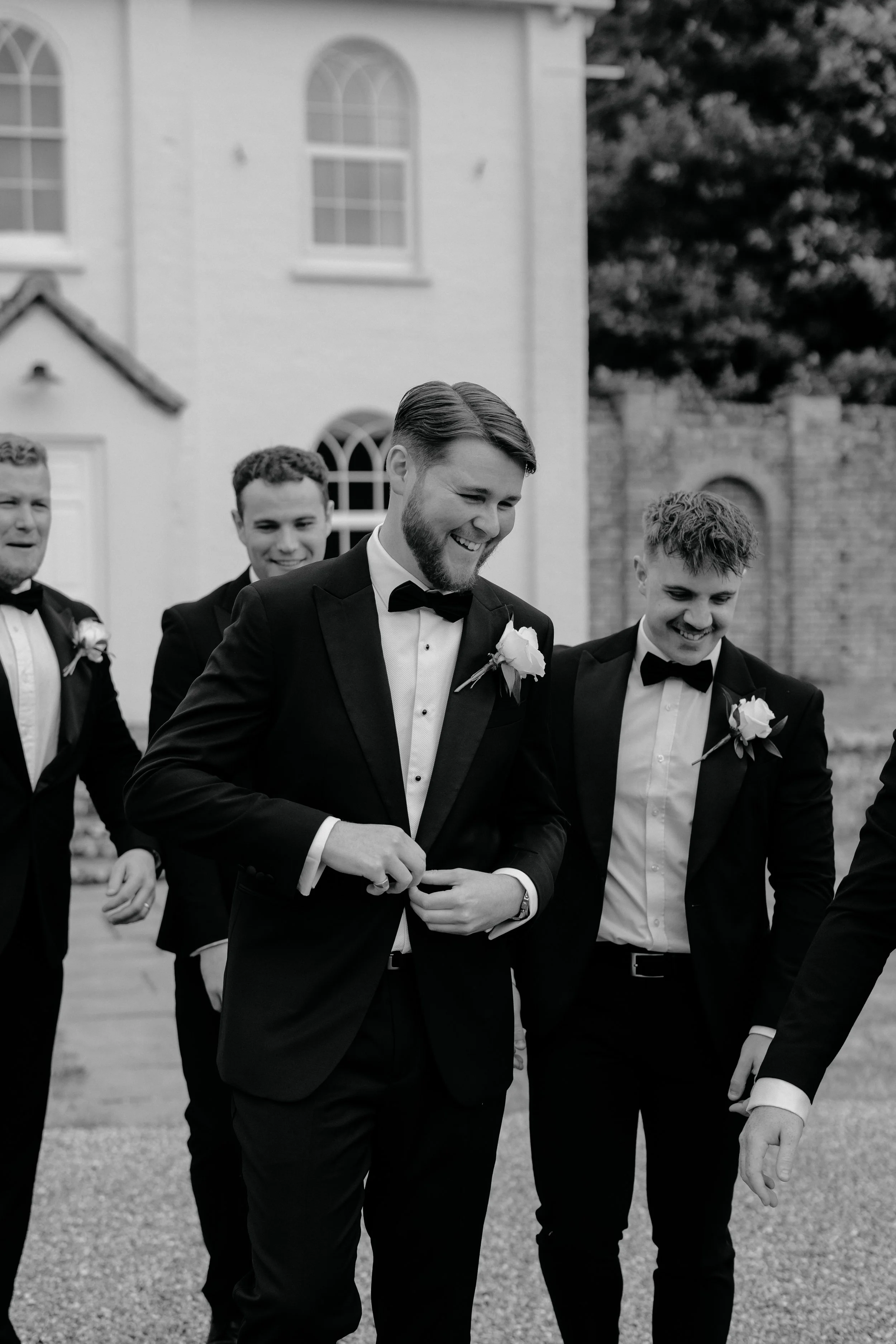 Black-and-white photo of four young men in tuxedos with bow ties and boutonnières, standing outdoors in front of a brick building with arched windows, smiling and looking down.