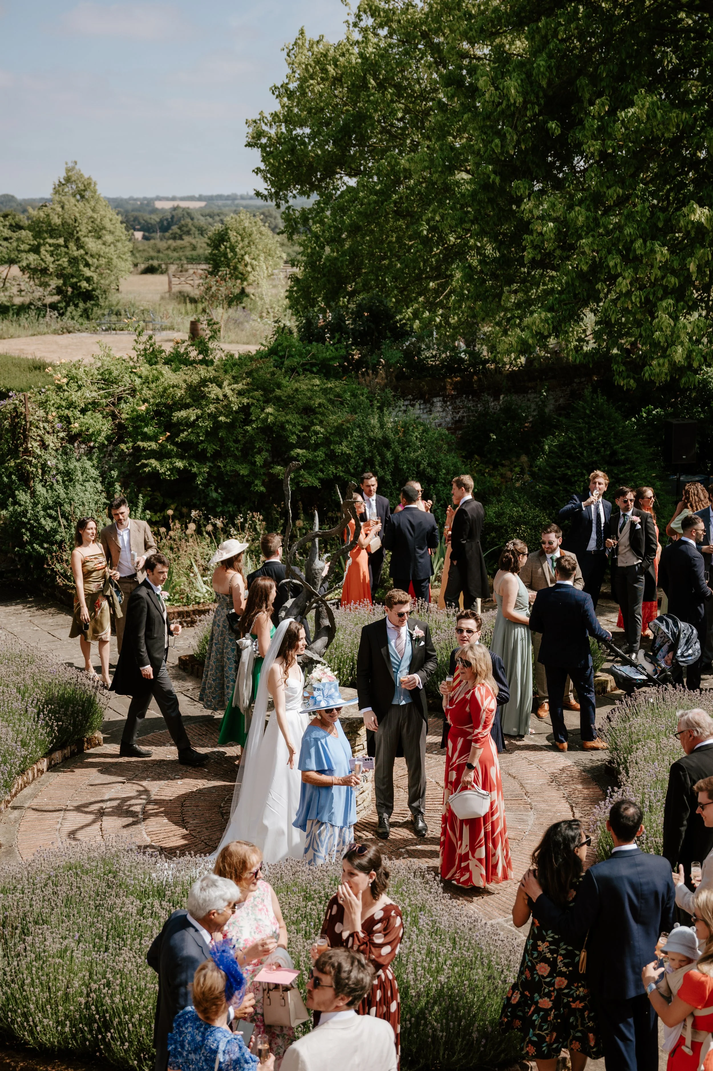 Scene of a garden wedding reception with guests mingling and socializing among flowering plants and trees on a sunny day.