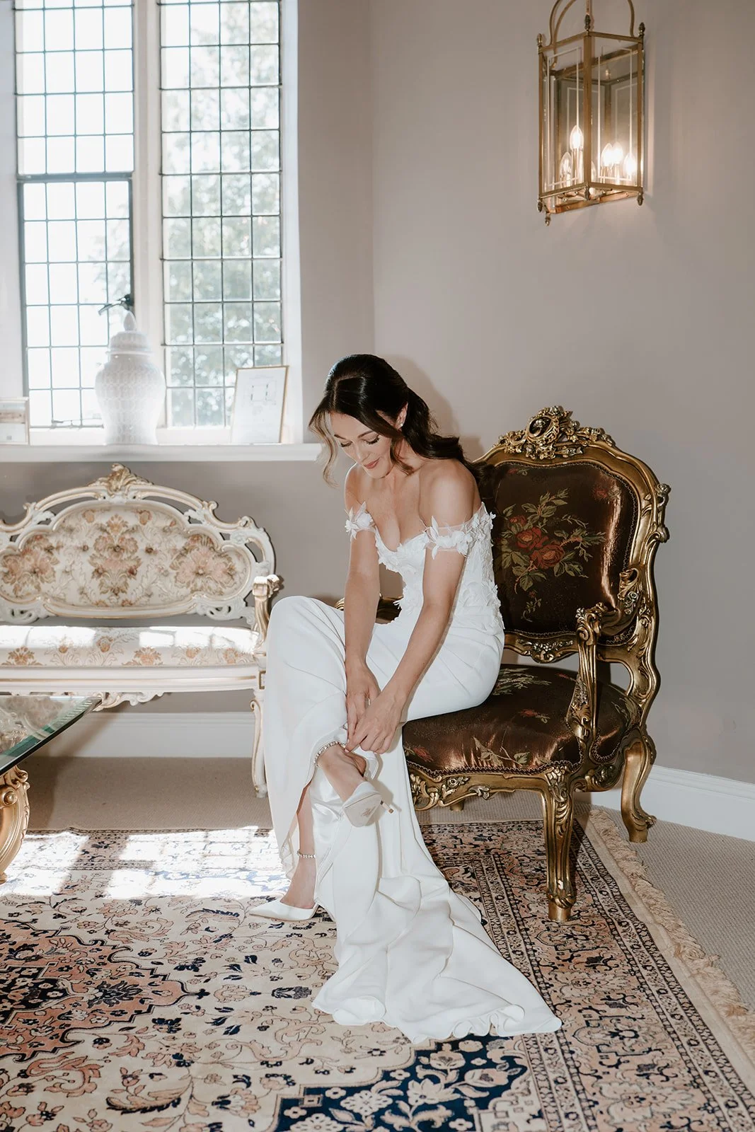 A bride in a white wedding gown is sitting on a vintage chair, adjusting her shoe in a decorated room with a floral couch, ornate gold furniture, and a chandelier.