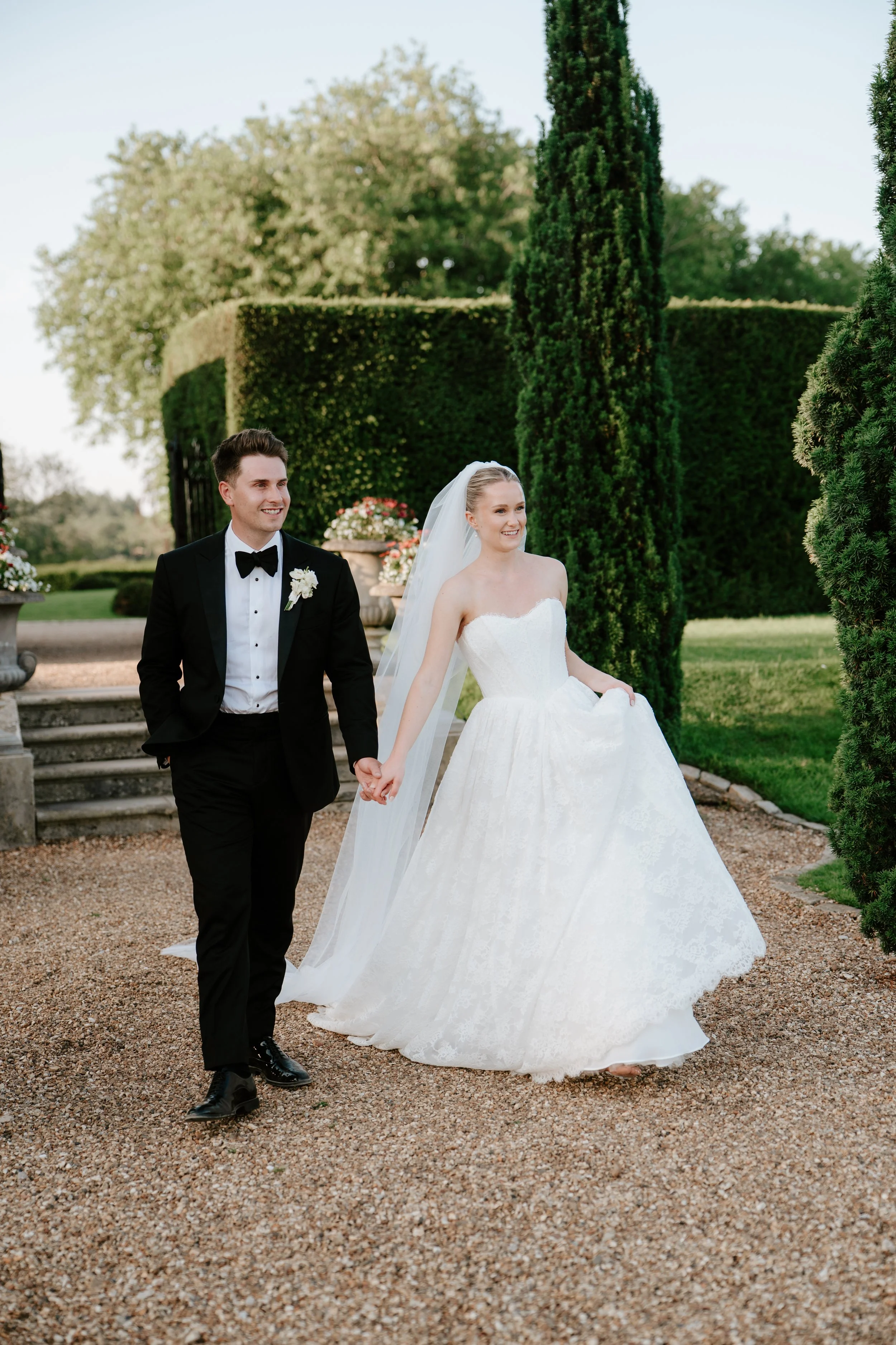 A newlywed couple walking hand in hand outdoors in a garden, with the groom wearing a black tuxedo and bow tie, and the bride in a white wedding dress and veil, surrounded by greenery and decorative flower pots.