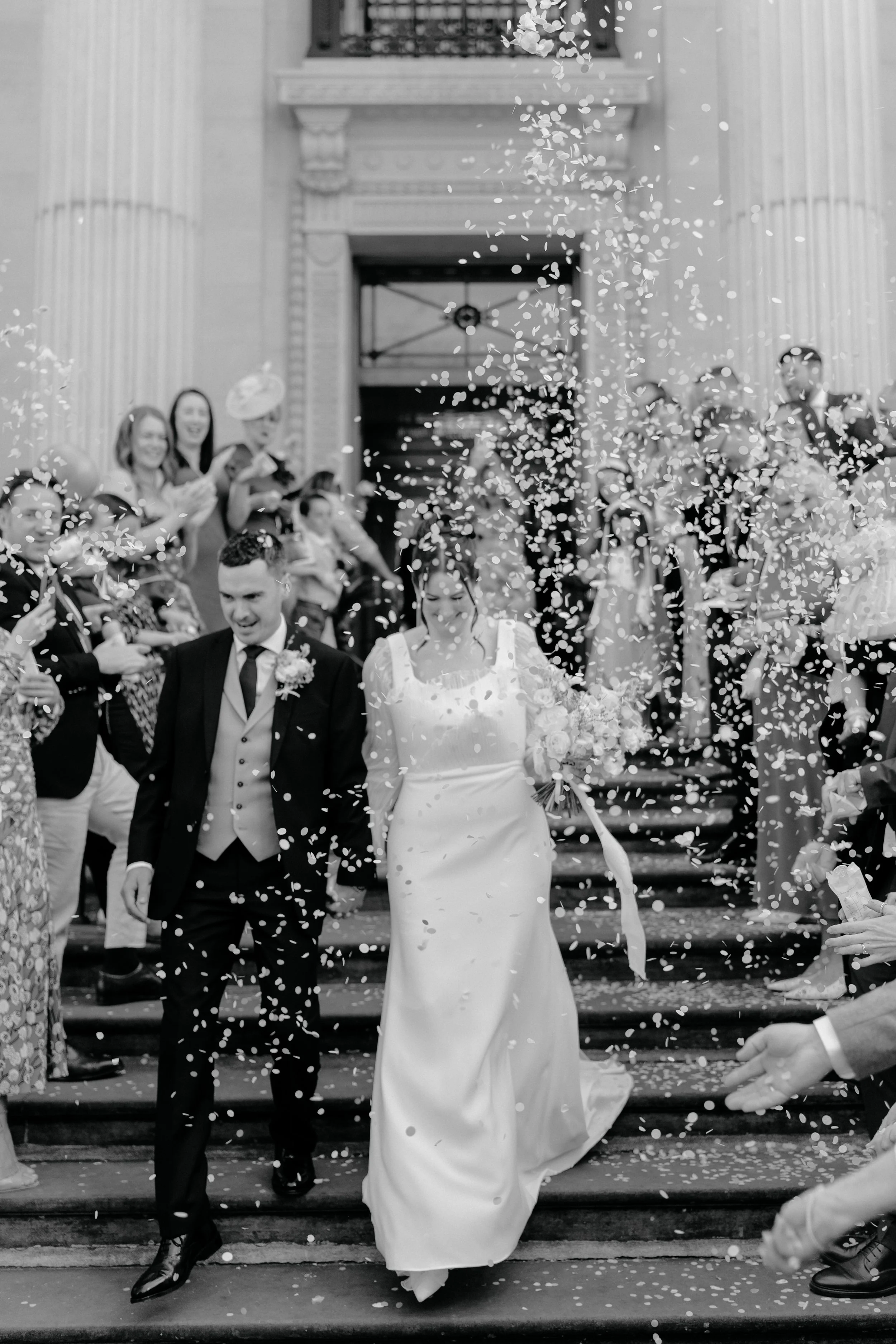 A wedding celebration with a bride and groom walking down the stairs surrounded by guests throwing confetti.