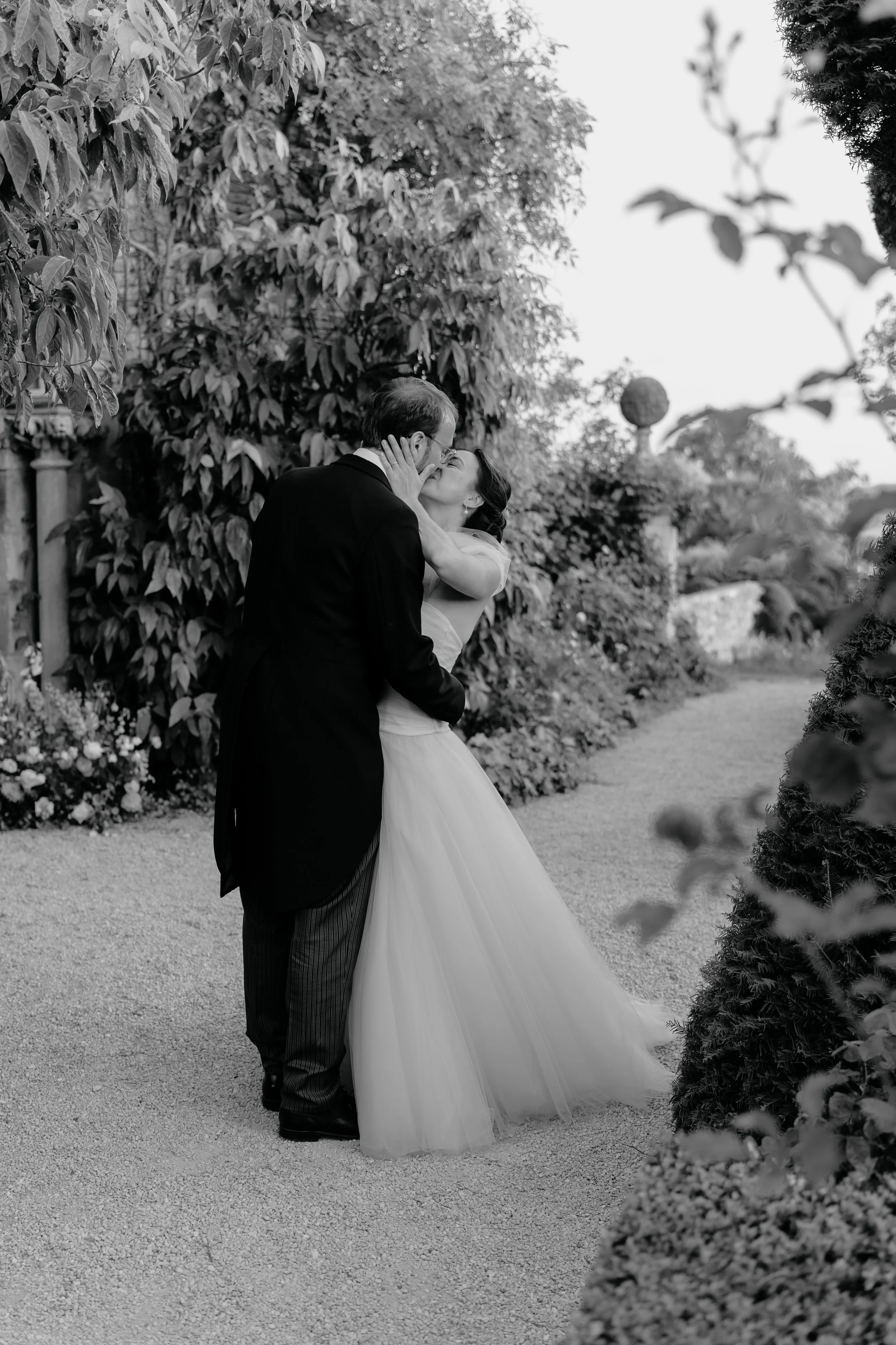 A black and white photo of a couple sharing a kiss outdoors, surrounded by trees and bushes.
