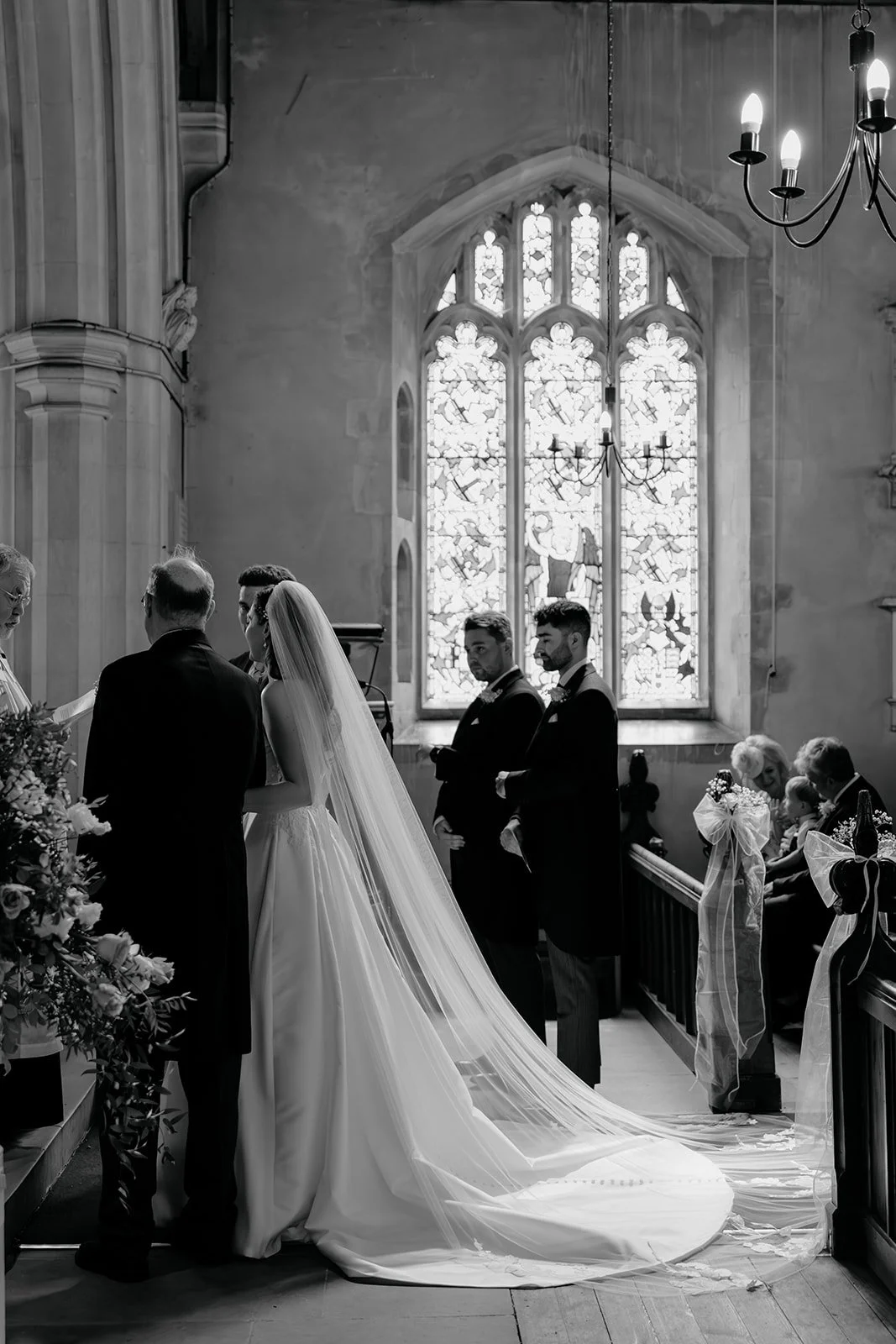 A wedding ceremony inside a church with a bride in a long white gown and veil, standing with a groom and an officiant. The church features a large stained glass window and hanging chandeliers, with guests seated along the pews observing the ceremony.