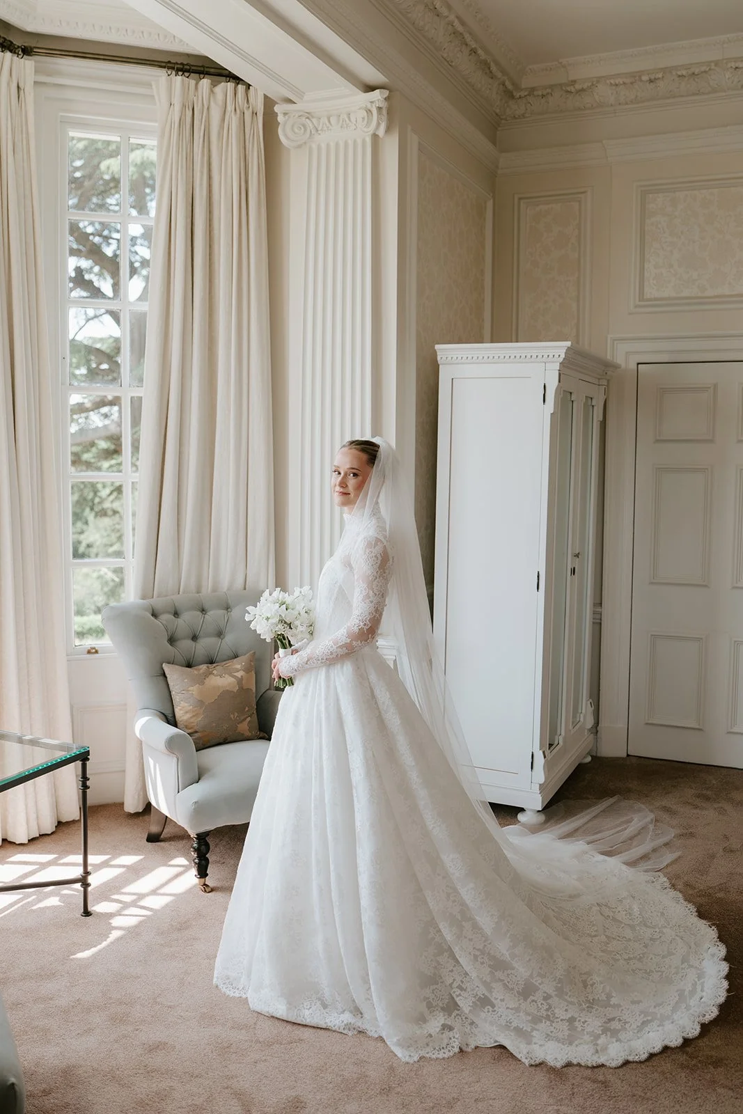 Bride in a white lace wedding dress standing in a well-lit room holding a bouquet of white flowers, with large windows, heavy curtains, and elegant molding in the background.