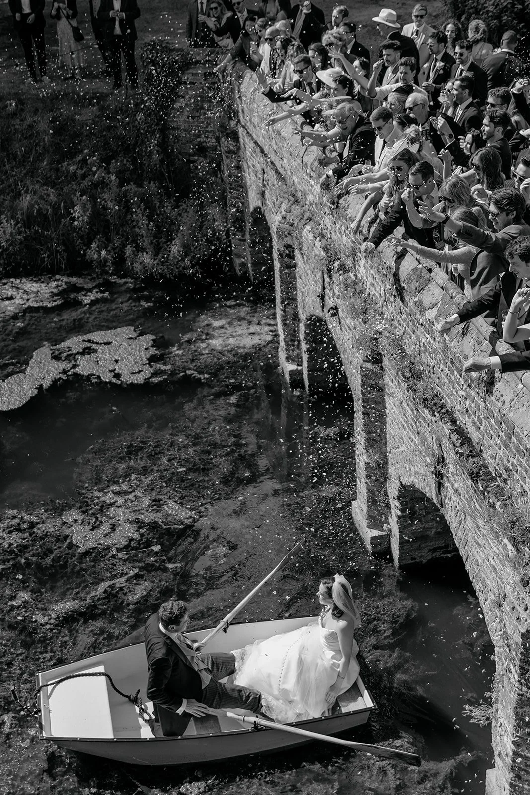 A bride and groom in a boat near a brick wall with a water fountain, with a large crowd of people leaning over the wall and throwing water on the fountain.