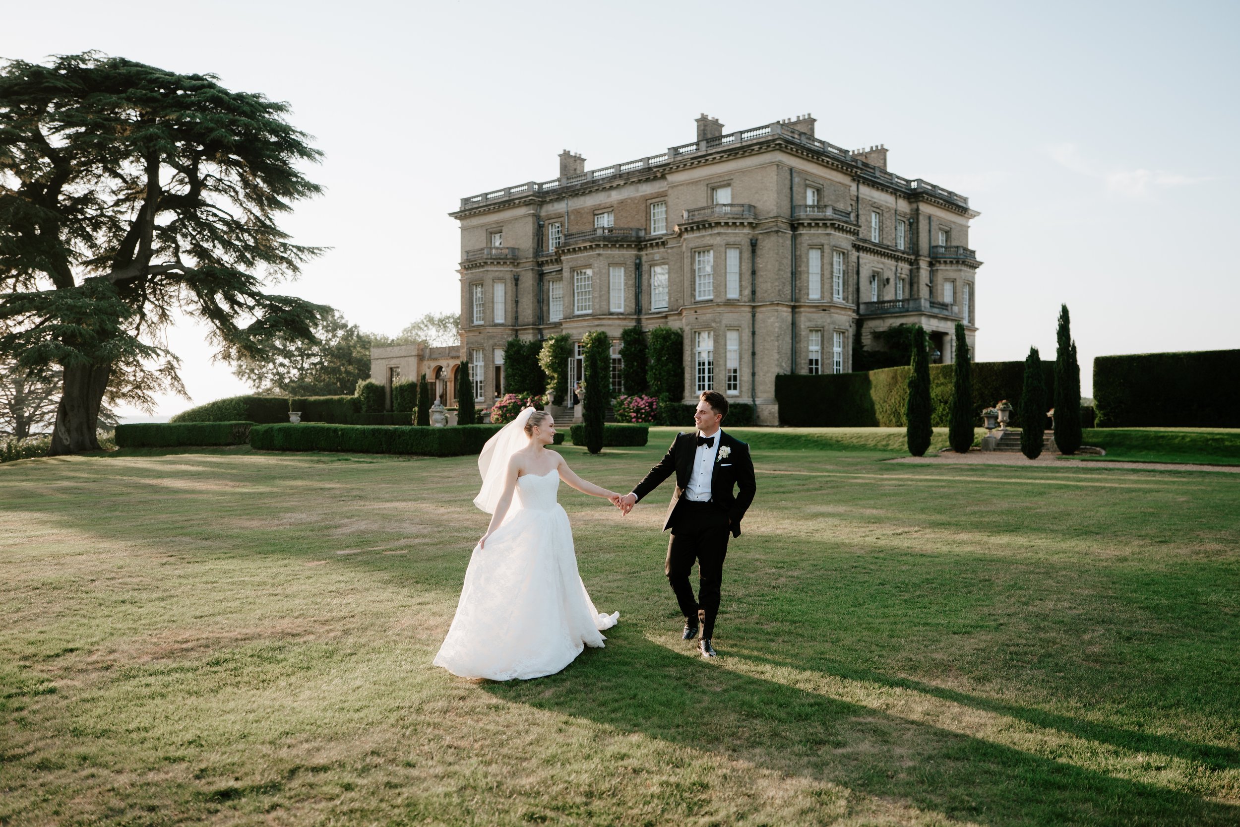 A bride and groom holding hands and dancing outdoors on a grassy lawn in front of a large historic mansion with tall trees and manicured bushes during sunset.