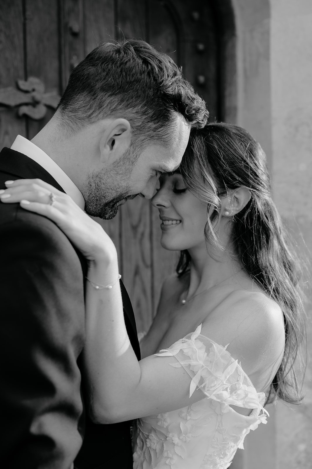 A black and white photo of a couple in love, with foreheads touching and eyes closed, smiling. The man is wearing a suit, and the woman is wearing an off-shoulder wedding dress with floral details.