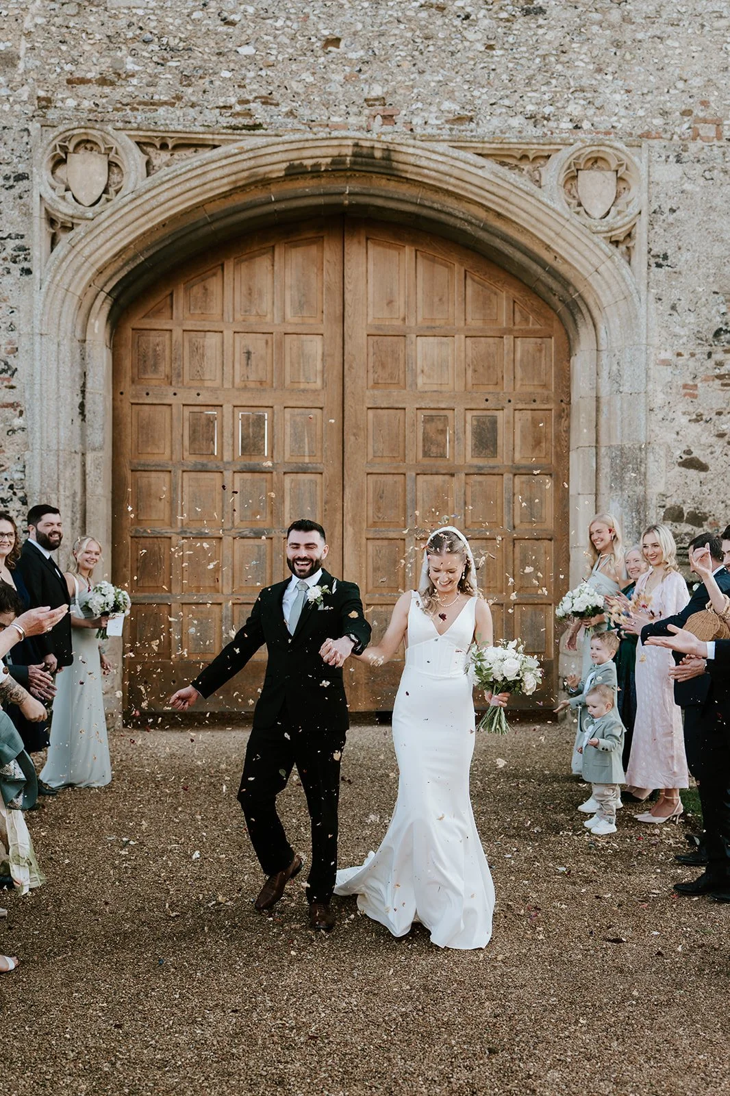 Bride and groom exiting church, surrounded by cheering guests, some throwing confetti, in front of large wooden door.