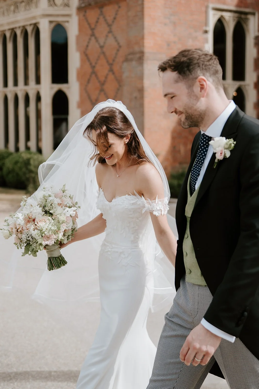 Bride and groom walking outside a brick church, smiling and holding hands. Bride in white wedding dress with a veil, holding a bouquet of flowers. Groom in black suit with boutonniere, smiling at bride.