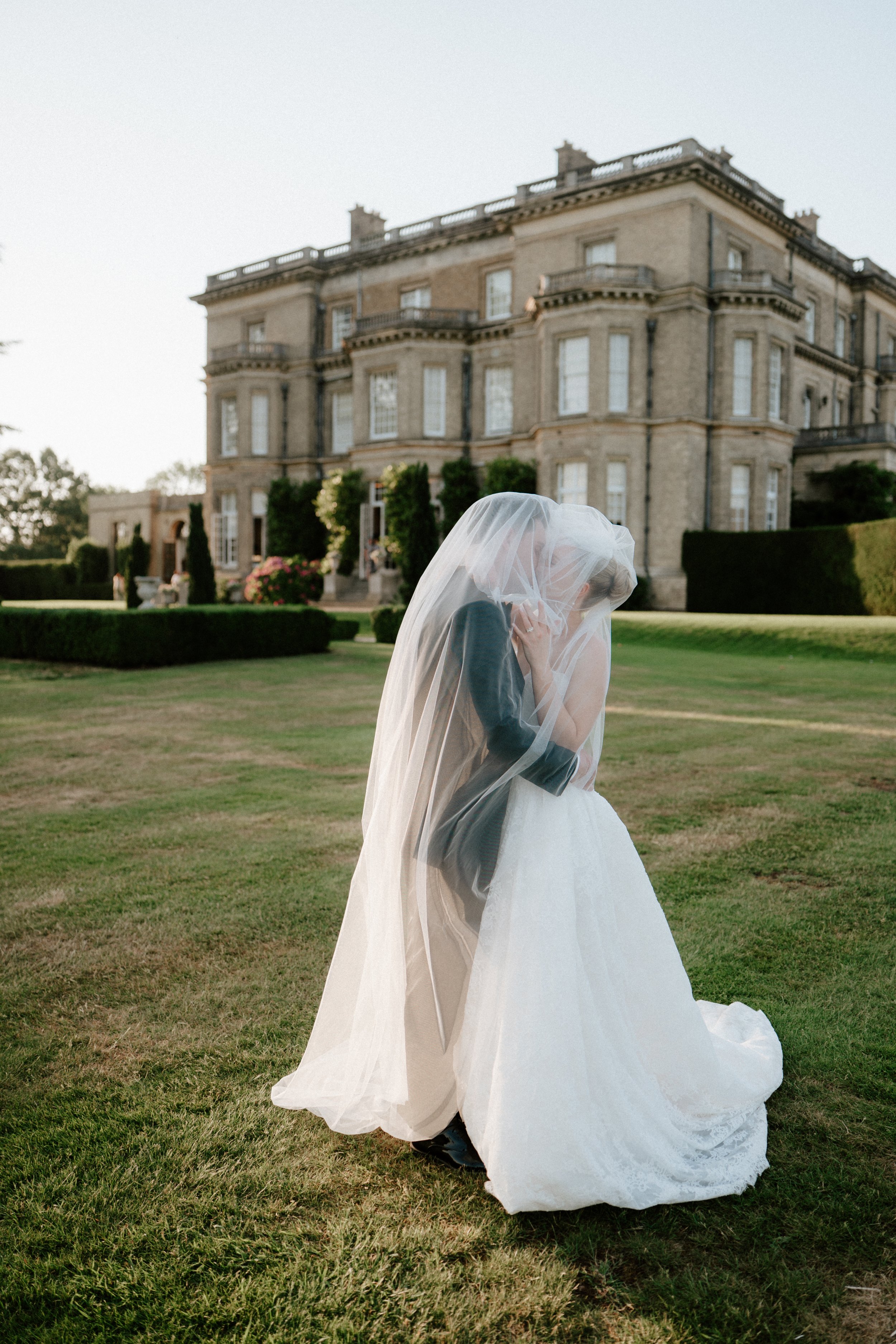 A bride and groom kissing under a wedding veil outside on a lawn in front of a large historic building with multiple windows and balconies.
