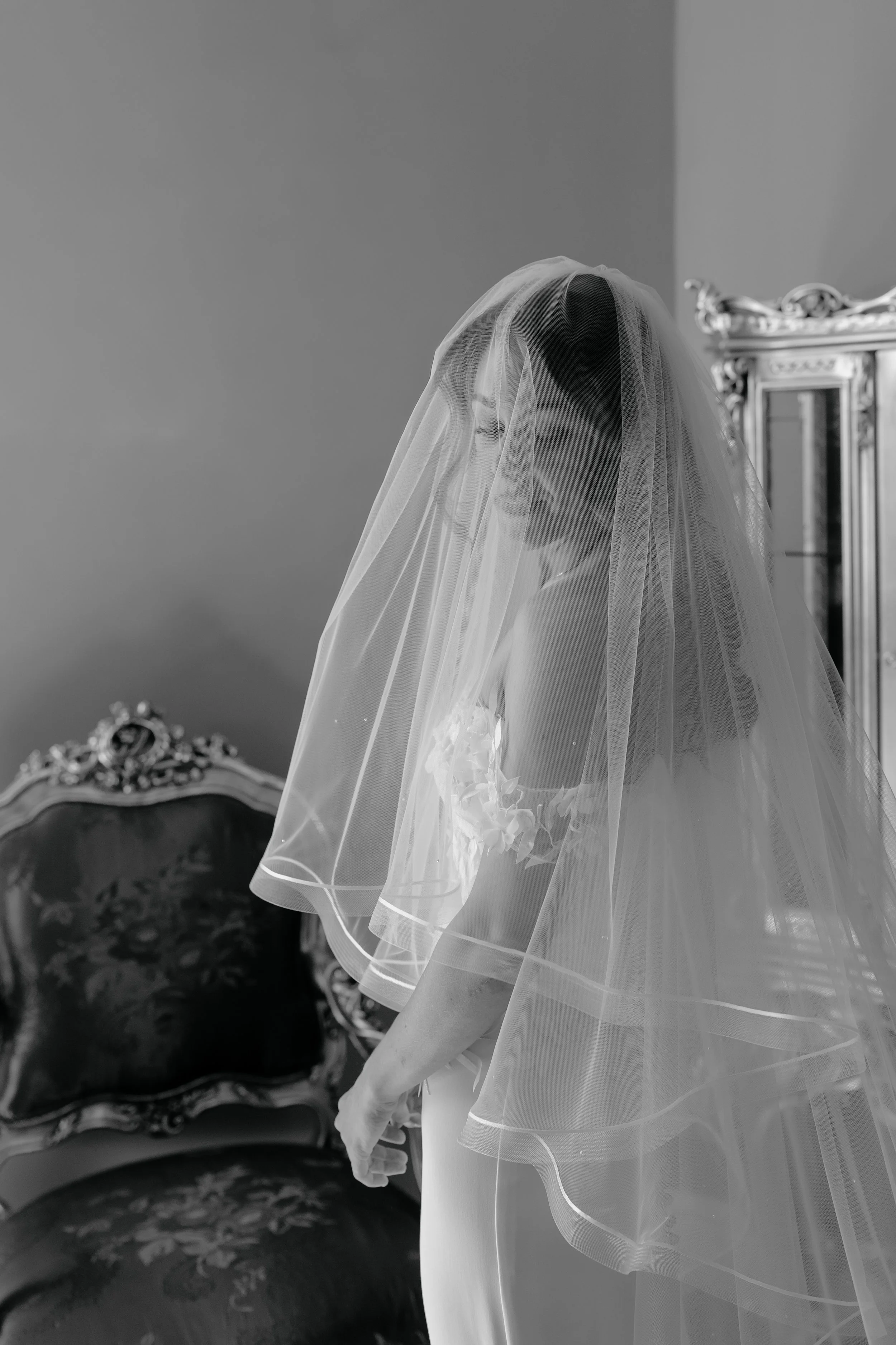 A woman in a wedding dress with a veil, standing indoors near an ornate mirror and vintage furniture, in black and white.