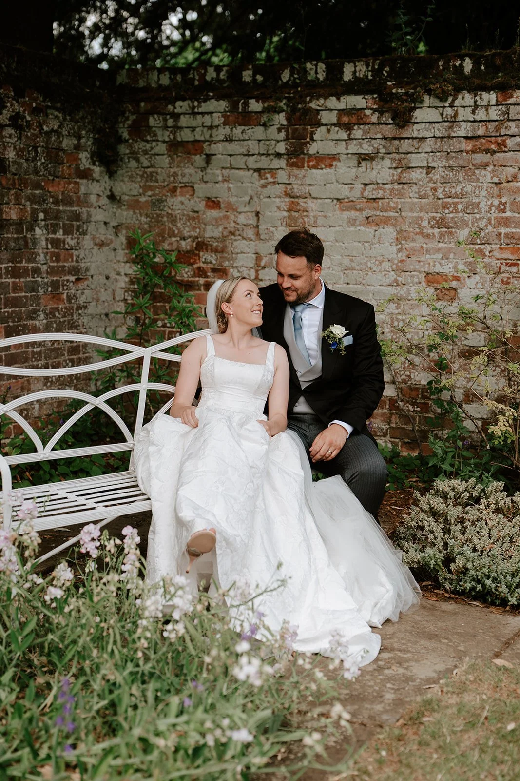 A bride and groom sitting on a white metal bench outdoors near a brick wall, sharing a moment of laughter and affection, surrounded by greenery and flowers.