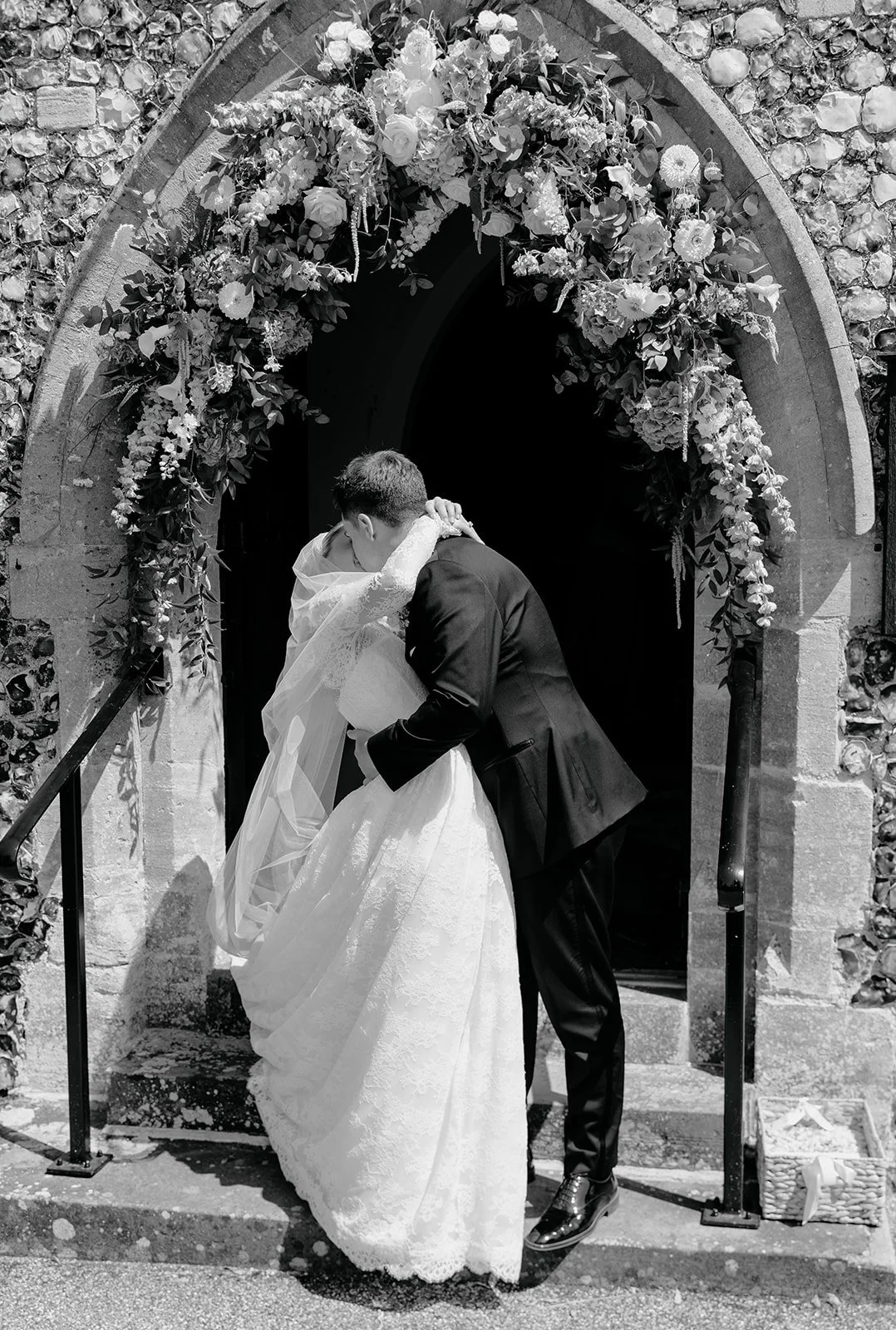 A black-and-white photo of a bride and groom sharing a kiss outside a church decorated with a floral arch.
