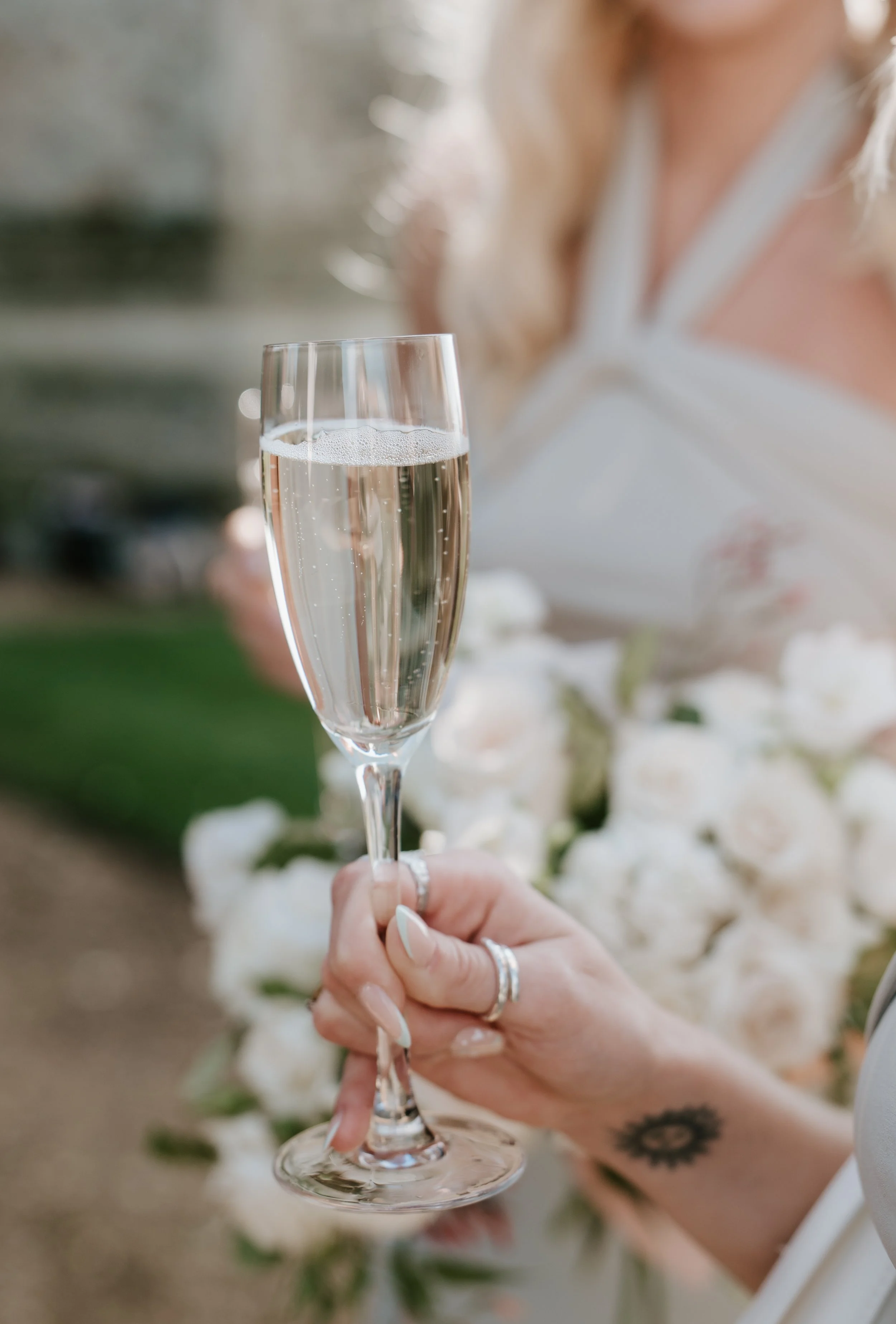 A hand holding a glass of champagne with a blurred person and floral decorations in the background.