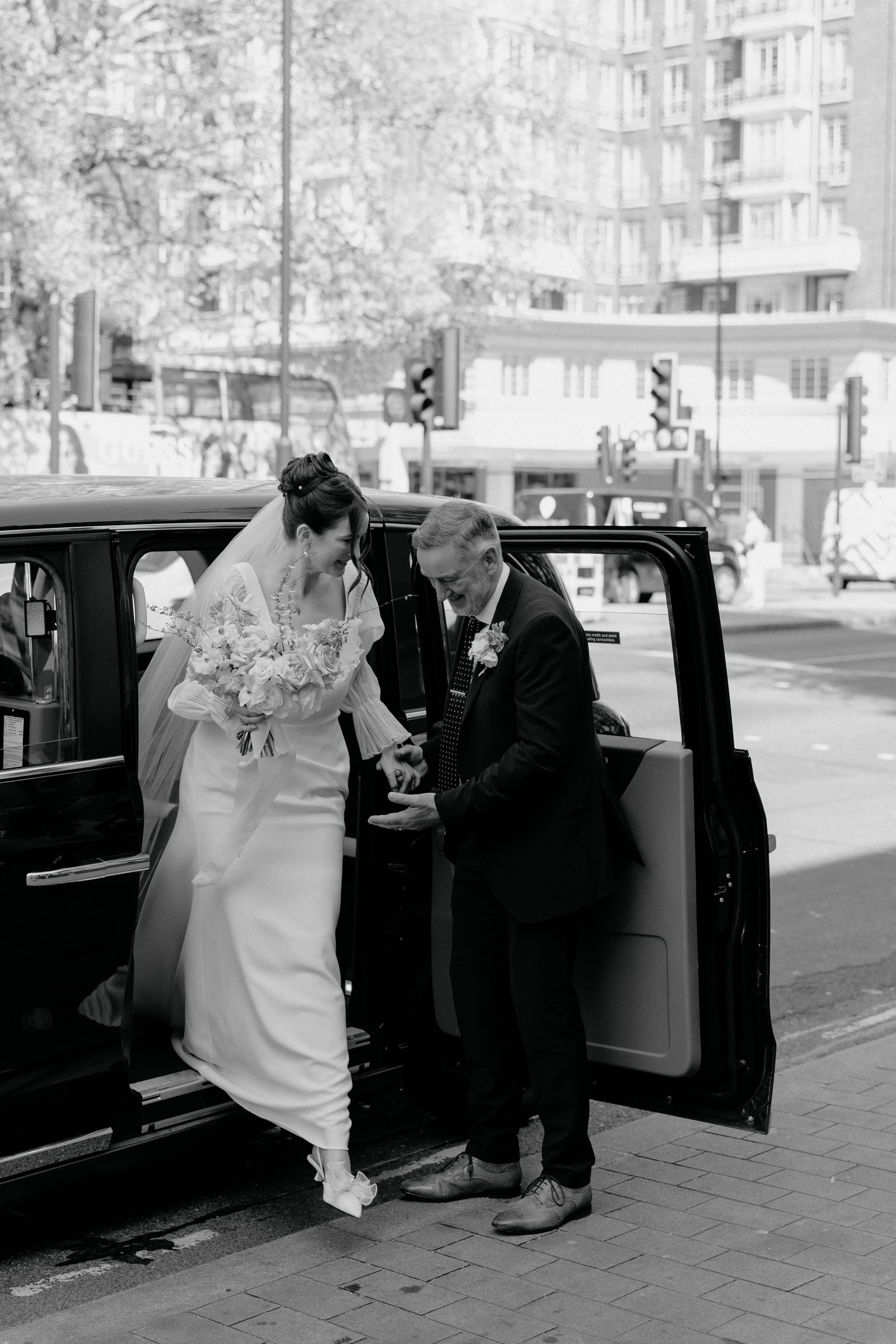 A bride in a white wedding gown and veil is stepping out of a black vehicle, assisted by an older man in a suit. She is holding a bouquet of flowers and looking at the man, smiling. The man is helping her out of the vehicle, on a city street with tall buildings and traffic lights in the background.