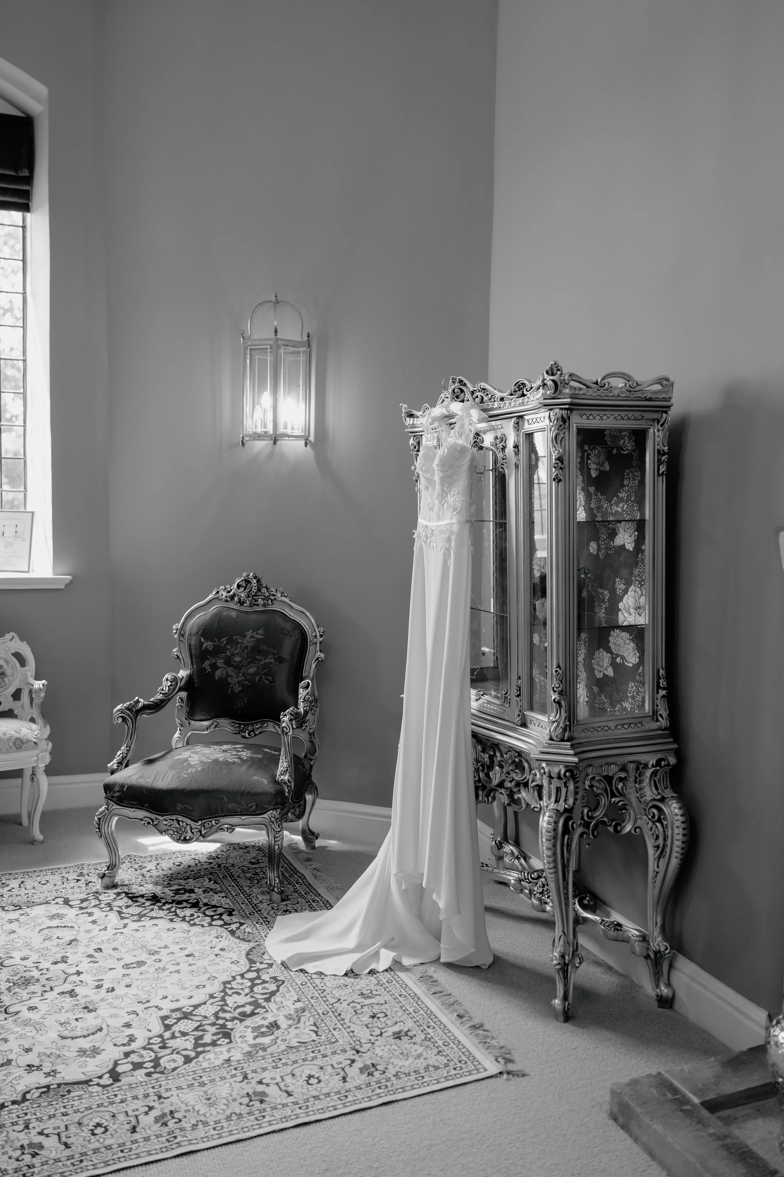 A vintage interior room with ornate furniture, including a high-backed chair, a glass display cabinet, and a wedding dress hanging on a stand, with a window, a wall-mounted light, and a patterned rug.