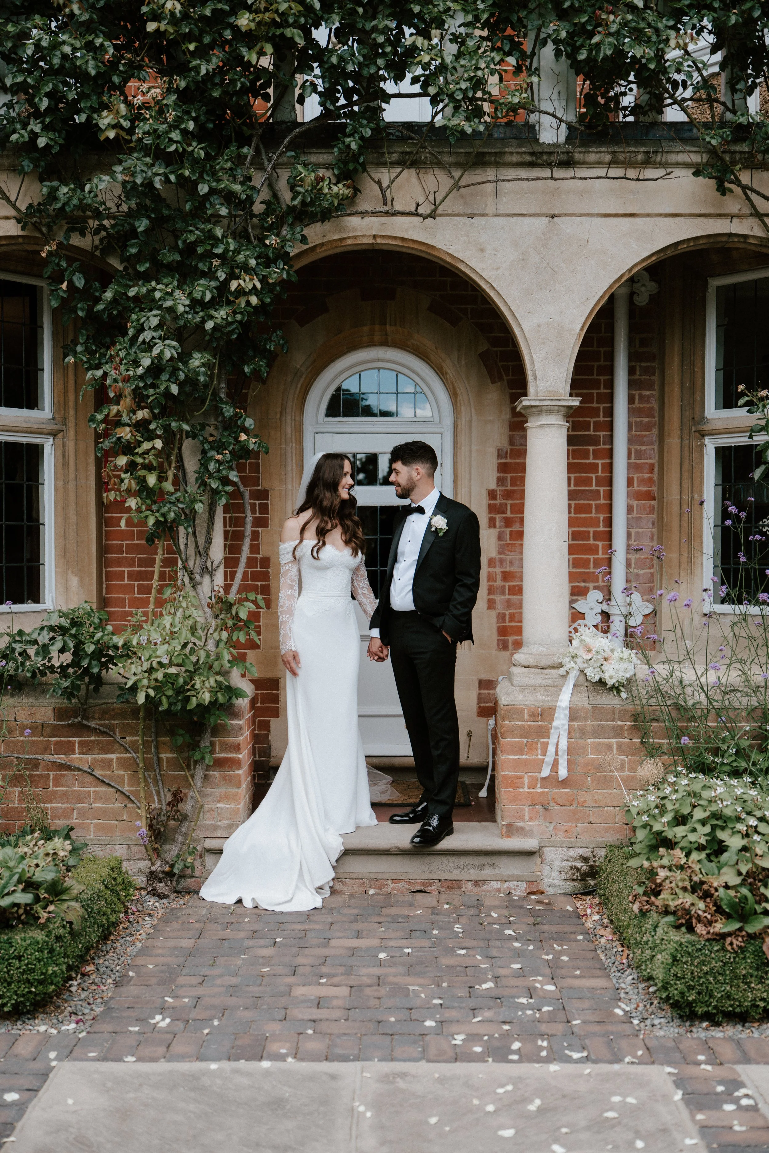 A bride and groom standing on the steps of a brick house with arched entrance, holding hands and looking at each other, surrounded by greenery and flowering plants.