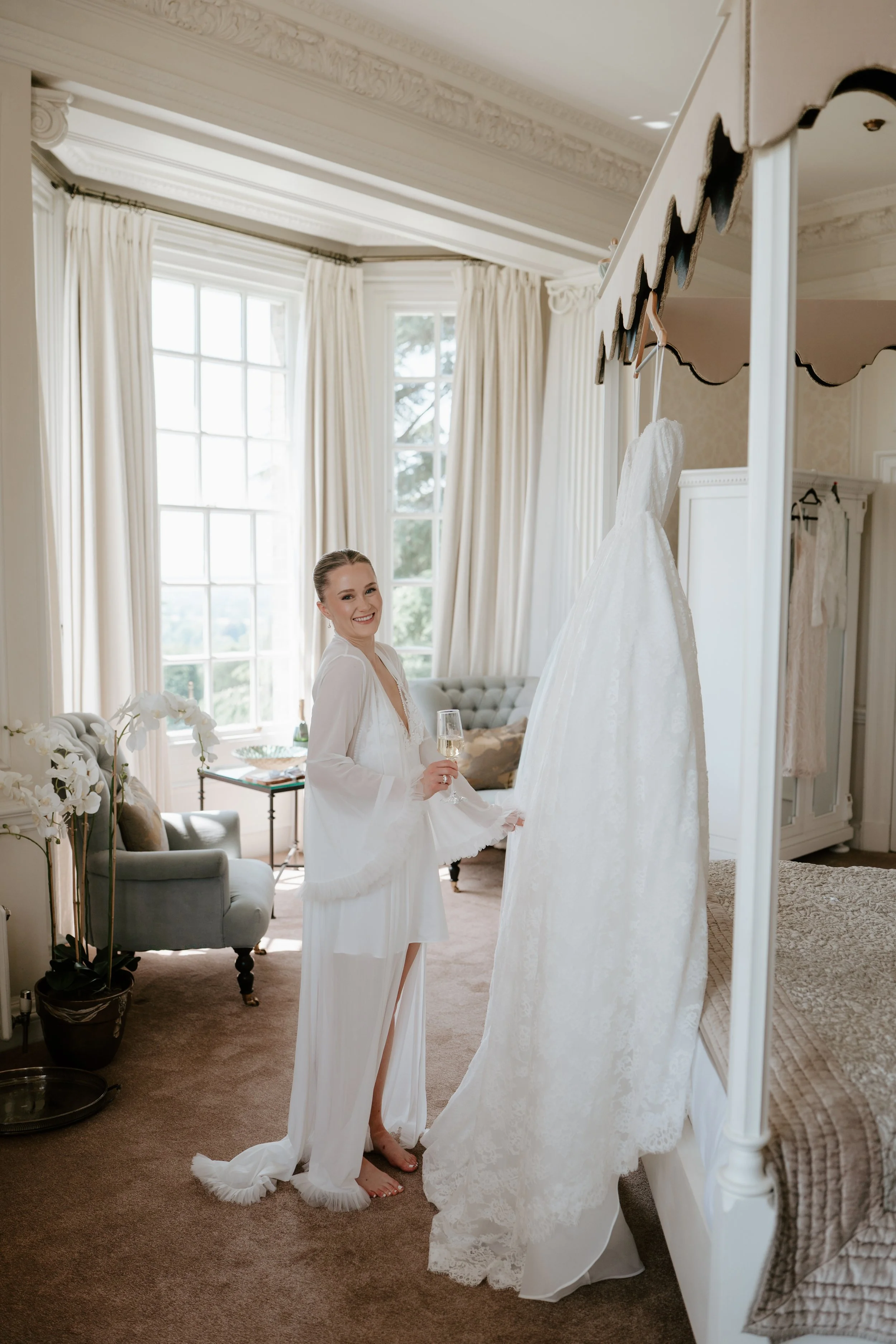 A woman in white dress smiling while holding a glass of champagne, standing in a bright, elegant room with large windows, curtains, and a wedding gown hanging on a mirror.